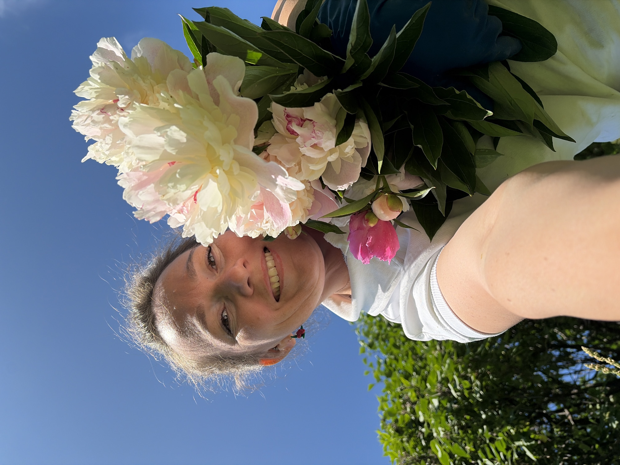 This is a vertical, low-angle, full-body shot of a woman holding a bouquet of peonies against a bright blue sky and green foliage. The woman is in the foreground, looking upwards at the camera with a wide smile, her face framed by the flowers. She is wearing a white hooded sweatshirt. Her arm and shoulder are visible, and her skin is tanned. The bouquet consists of large, creamy white and pale pink peonies, with some smaller pink buds. The lighting suggests it is daytime, likely sunny. The overall mood is joyful and vibrant, with the flowers adding a touch of natural beauty and color. The background includes lush green leaves and branches of trees, and the clear blue sky indicates fair weather.
