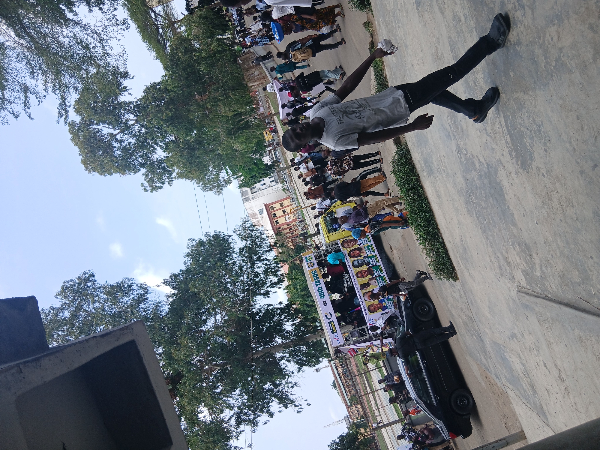 This image captures an outdoor daytime scene of a public gathering or rally, likely related to a student election or campaign, set in Lagos, Nigeria.
In the foreground, a man wearing a grey t-shirt with "PALMTREE" printed on it and black pants stands, holding a crumpled white object in his right hand. He is partially turned away from the camera.
The midground features a crowd of people, some standing and others seated, gathered around a temporary stage or setup. A large banner, central to the event, displays multiple faces, presumably of candidates, and campaign text. Visible text on the banner includes "VOTE Catalyst", "Team BASSEY", and "46th YASUG". A clear slogan states: "Bridging the gap between the management and the students'". A black car is parked near the banner.
The background shows more individuals, along with several buildings and numerous lush green trees that line the area. The sky is bright blue with scattered white clouds, indicating clear weather. The ground is paved, and there are patches of greenery.
The activity suggests an organized event, possibly a political rally or campaign meet-and-greet within a university or college setting, given the "YASUG" and "students" references. The overall atmosphere appears orderly and engaged.