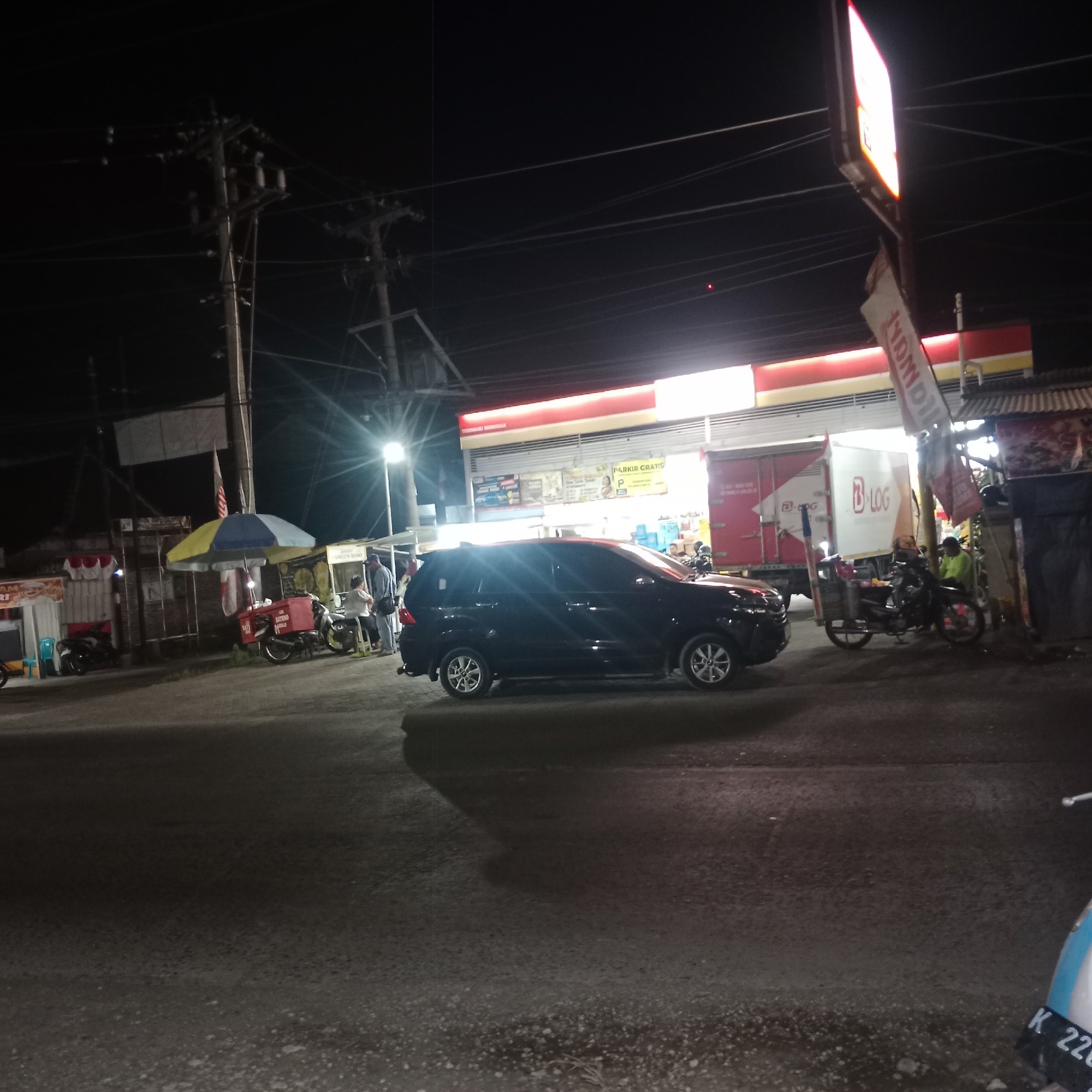 The image captures a nighttime scene in front of a minimarket in Tegowanu Kulon, Indonesia, likely a 7-Eleven or similar convenience store, indicated by the red and white branding and the "PARKIR GRATIS" (Free Parking) sign. The foreground features a black SUV parked on the asphalt road, with its headlights off. To the left, a food stall with a blue and yellow umbrella is visible, with a motorcycle parked nearby. Further to the left, another food stall with red and white awnings is present. In the background, a white and red delivery truck with "B LOG" branding is parked. Several motorcycles are parked throughout the scene, and a few individuals are standing or sitting near the stalls and the truck. Power lines and poles crisscross the dark sky above, illuminated by streetlights and the bright signage of the minimarket. The overall atmosphere is that of a busy commercial area at night.