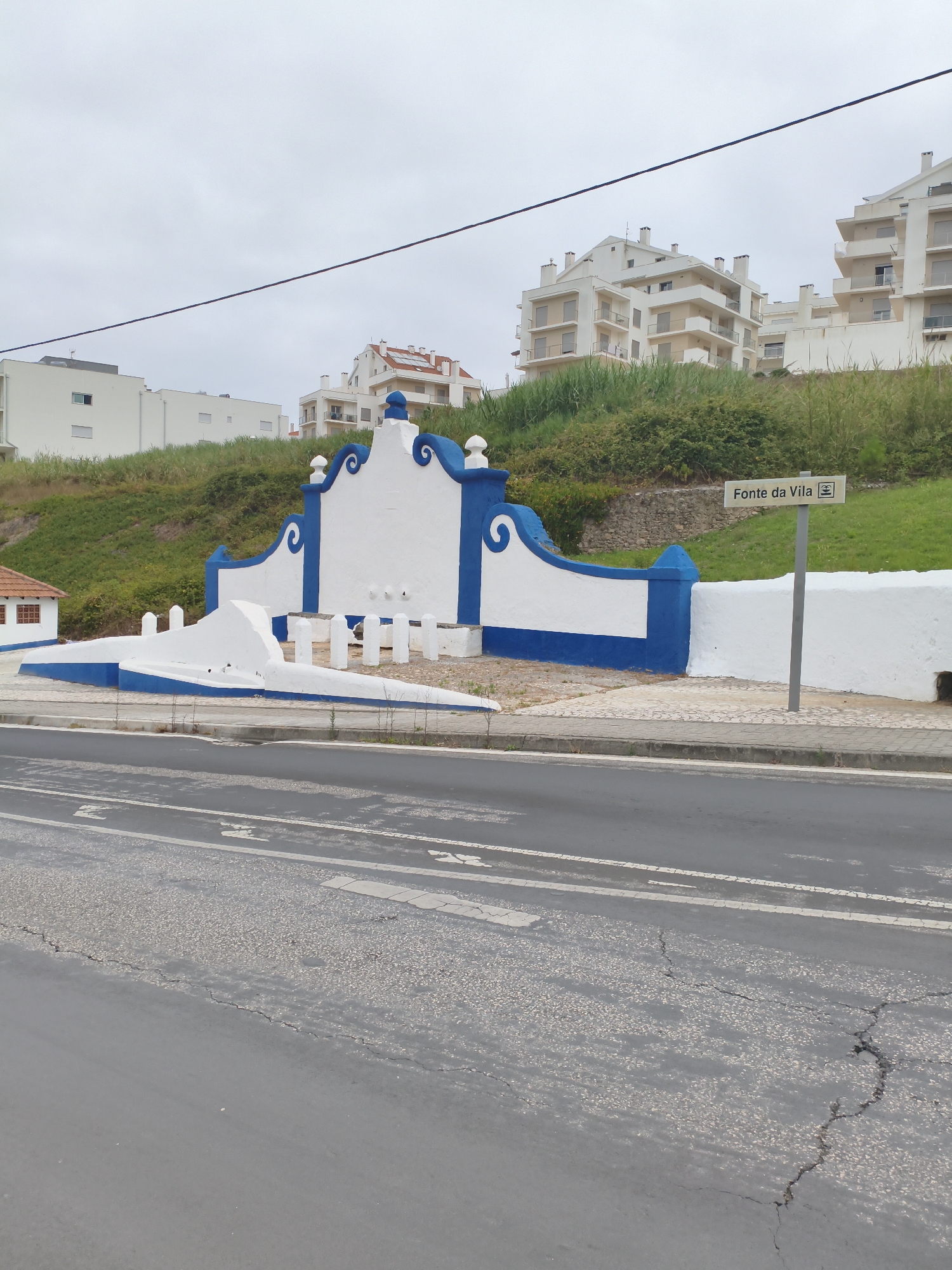 The image captures a street scene in Nazaré, Portugal. In the foreground, a gray asphalt road with visible cracks and white lane markings stretches across the frame. To the right, a sidewalk with light-colored paving stones leads to a street sign with text in Portuguese, "Fonte da Vila", and a symbol that appears to represent a fountain.
Behind the sidewalk is a decorative white and blue structure that appears to be a public fountain or monument, with curved blue accents and a central white section. This structure is situated on a grassy incline. Above the fountain, several multi-story white residential buildings with beige trim are visible, overlooking the scene. The sky is overcast and a dark cable is stretched across the upper portion of the image. A small, red-roofed building with brown window frames is partially visible to the left of the fountain. No people are present in the image.