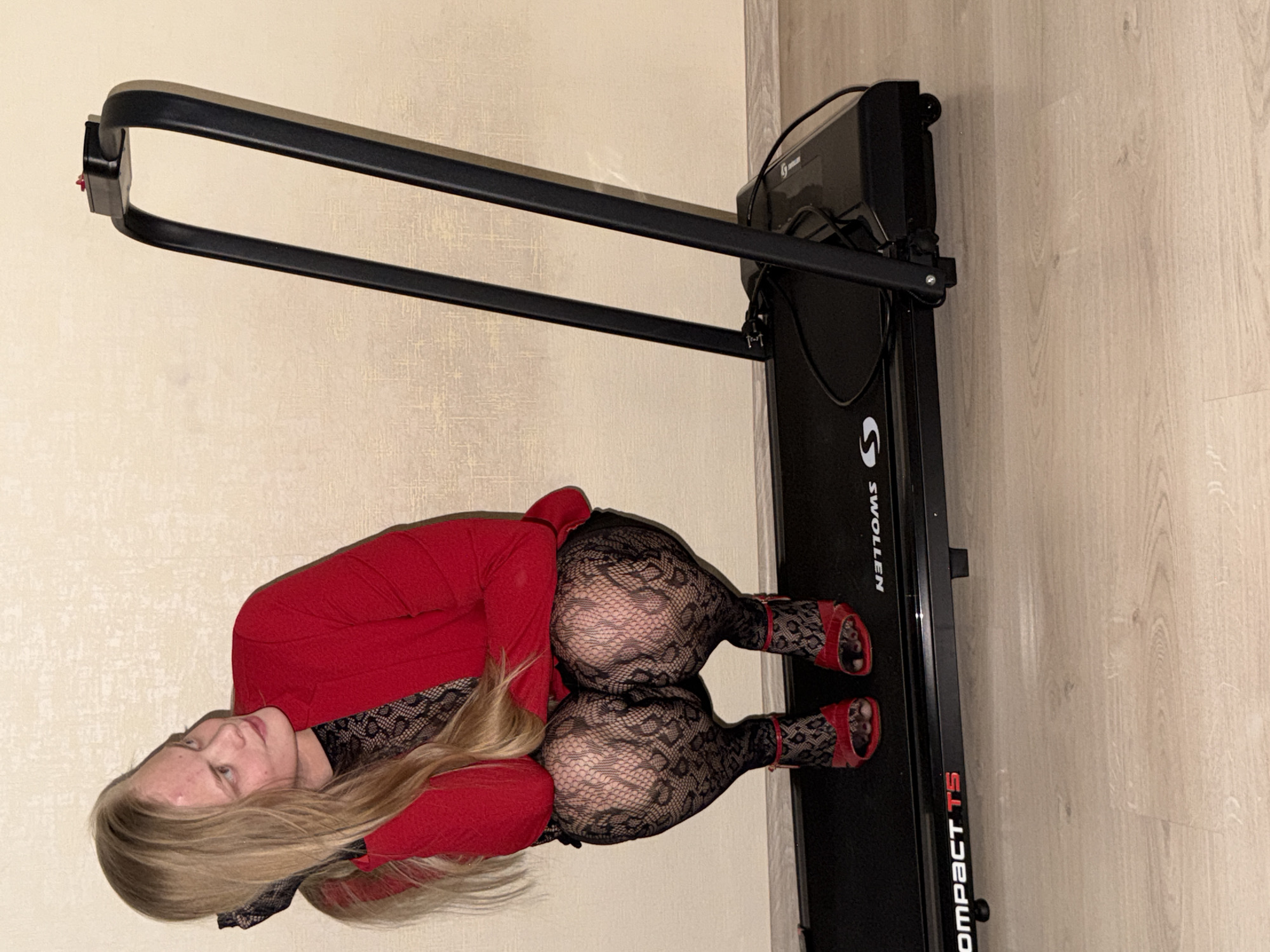 An indoor, portrait-oriented photograph depicts a young adult posing on a black treadmill. The image appears to be rotated 90 degrees clockwise. The subject has long blonde hair and is wearing a red long-sleeved top, black lace-patterned leggings, and red open-toed high-heeled sandals. She is lying on her stomach on the treadmill, with her head lifted and looking directly at the camera, displaying a neutral expression.
The treadmill itself is black, with the brand name "SWOLLEN" and model "COMPACT T5" visible on its frame. The handles and control panel of the exercise equipment are also visible. The background consists of a light-colored, textured wall and light-toned wooden flooring, typical of a modern home or apartment interior. The scene is illuminated by artificial lighting. This is a posed shot, focusing on the individual and her attire rather than an exercise activity, set in what is understood to be Izhevsk, Russia.