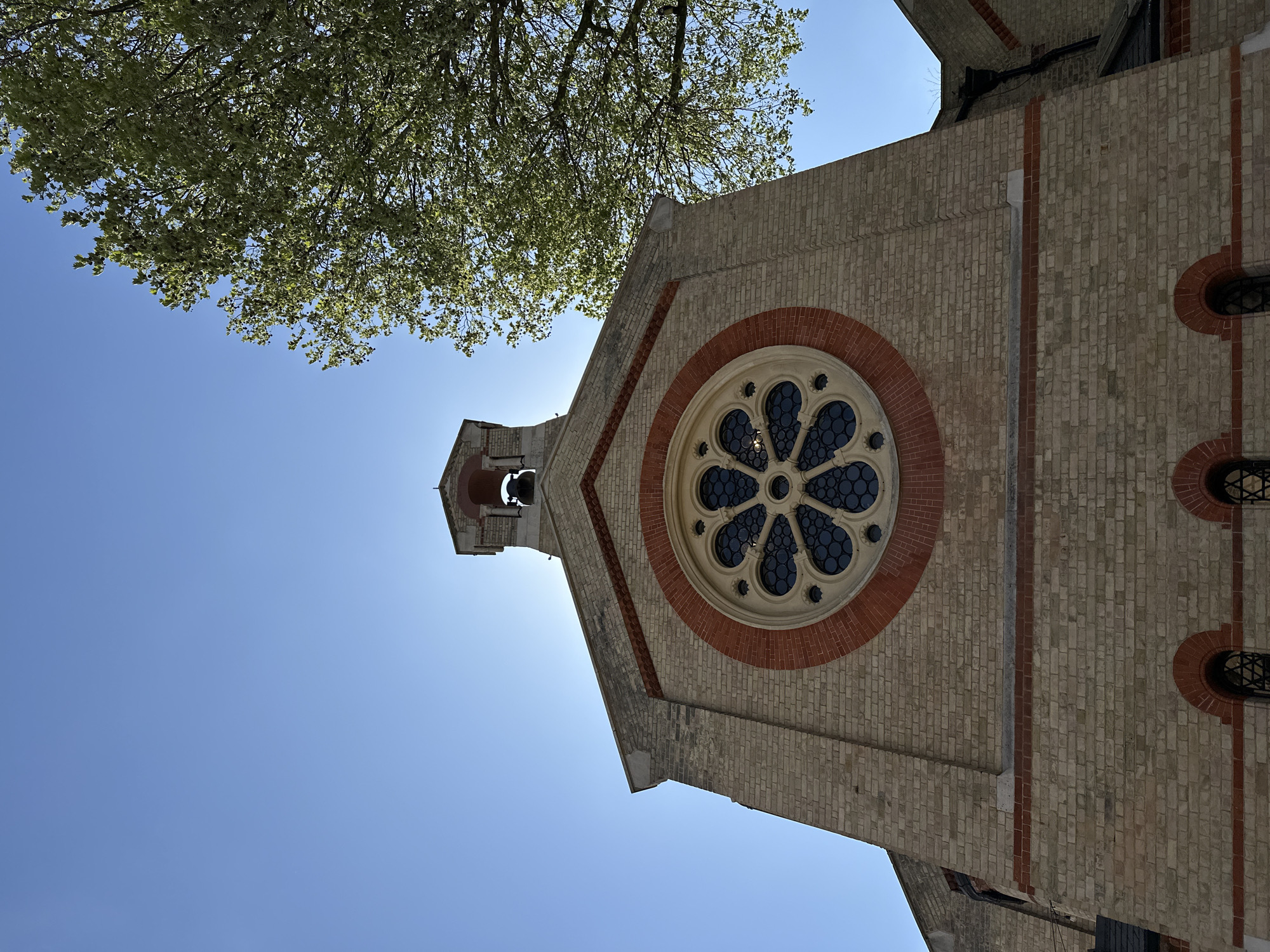 The image captures a low-angle perspective of a brick building, likely a church, viewed upwards against a clear blue sky. The photograph is oriented with the building occupying the right and upper portions, while the sky extends across the left and bottom.
The building features light-toned bricks with striking red brick details used for decorative bands and arches. A prominent circular rose window, characterized by intricate dark tracery, is centrally located on the main facade and framed by the contrasting red brickwork. Above and to the left of this window, a small belfry or bell tower is visible, housing a bell and silhouetted against the bright backdrop.
A canopy of green tree foliage overhangs the upper left quadrant of the frame, with individual leaves appearing illuminated by sunlight. The sky is a vibrant, cloudless blue, indicating clear and sunny weather. Strong backlighting suggests it is bright daytime, likely mid-day or early afternoon, with the sun positioned behind the structure. No people, specific activities, or visible text are present in the image. The architectural style points to a historical or religious structure in London, United Kingdom.