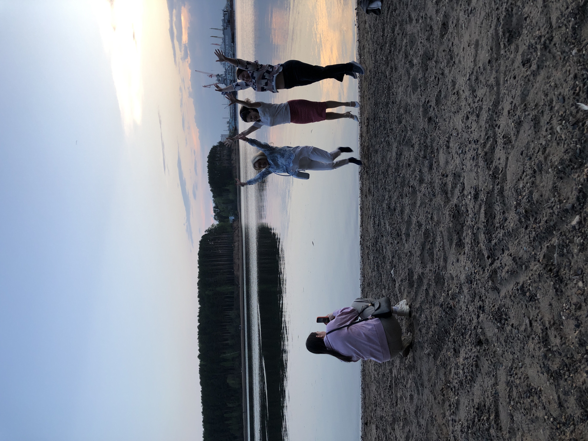 The image shows a beach scene at sunset in what is likely Chaikovsky, Russia, given the prompt's context. Three individuals, positioned horizontally one above the other, are mimicking a flying posture with arms outstretched. A fourth person, likely the photographer, is crouched nearby and taking a picture with a smartphone. The background consists of a body of water, probably the Kama River, and a line of trees along the opposite shore. The sky is softly lit with the colors of the setting sun, and industrial structures (cranes) are visible in the distance. The foreground is a sandy beach.