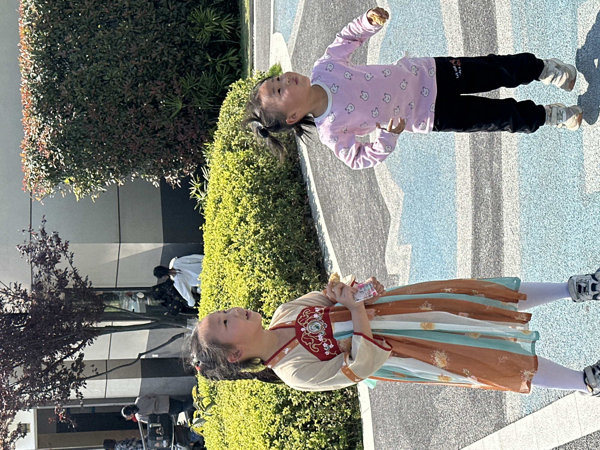 Two young girls are standing on a paved surface with a blue and gray pattern in an outdoor setting. The girl on the left is wearing a traditional Chinese dress with earth tones and an ornate red floral pattern on the bodice. She is holding a small clear container. The girl on the right is wearing a pink long-sleeved shirt with cartoon bunnies and black pants. She is holding a small round object. Both girls have their hair pulled back in pigtails and are looking upward. Green hedges and trees are visible in the background, along with a building with a modern architectural style. Some people are sitting down in the background near the building. The location is identified as Jiangjin, China.
