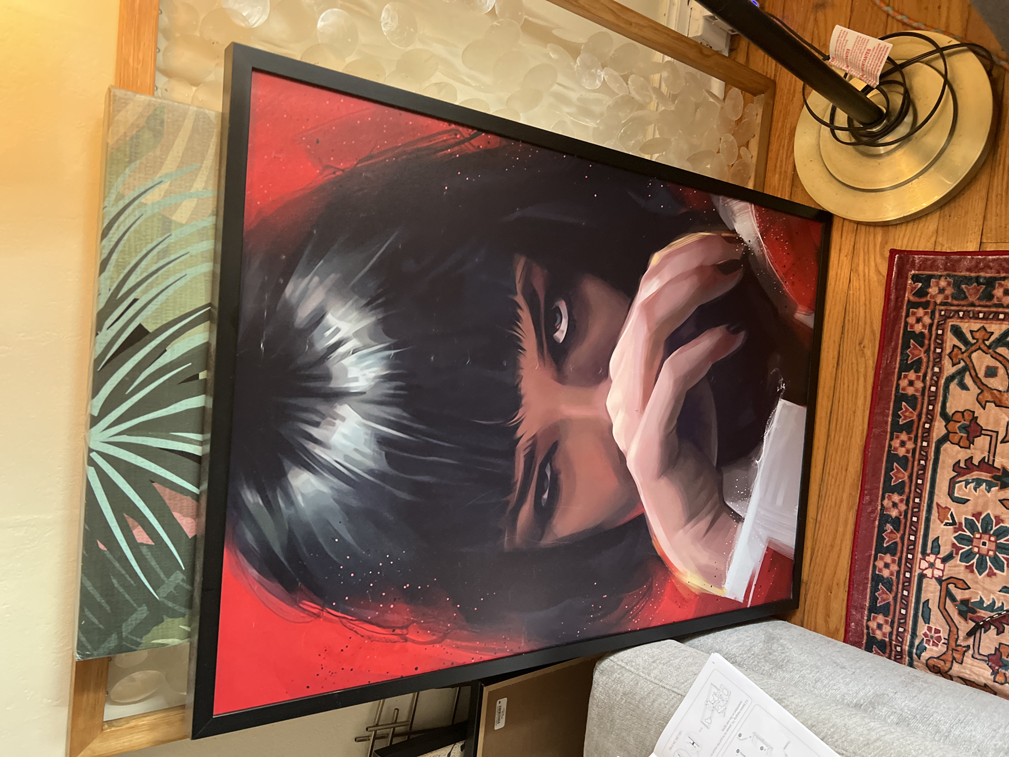 A high-angle shot depicts a framed portrait of Uma Thurman from Pulp Fiction with a bright red background and black hair, sitting askew on a gray upholstered surface. Next to the portrait are other items: a canvas painting with a green fern print, a gold-colored floor lamp, and a small carpet with a patterned design. The scene appears to be indoors, possibly in a home. The location is described as City of Kingston, United States of America.