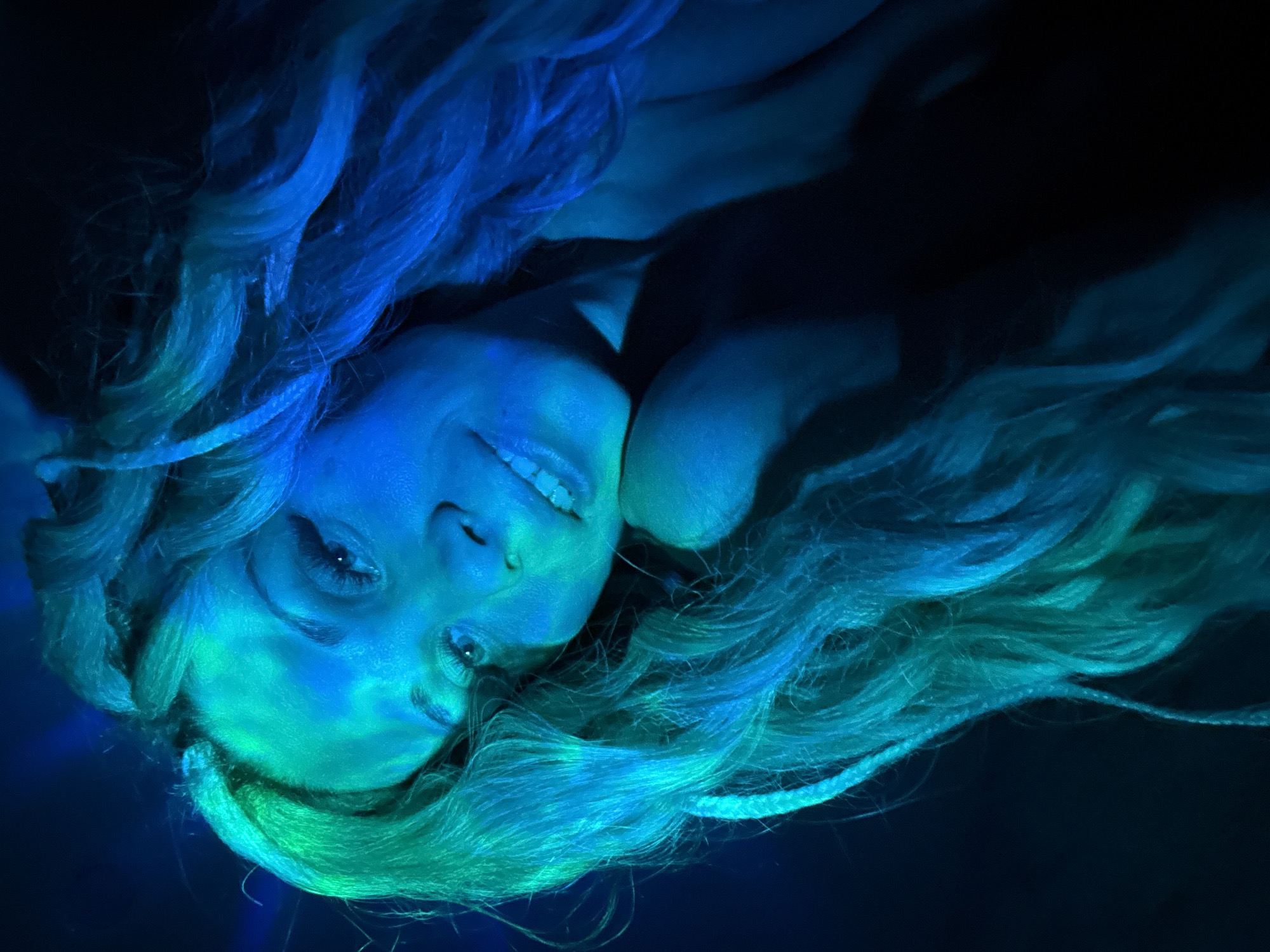 A woman with blonde hair and fair skin is lying down and looking up at the camera. She is smiling. Her hair is spread around her head and shoulders. The lighting is blue and green. The location is unknown and there is no evidence to suggest this is Rusanovo, Russia.