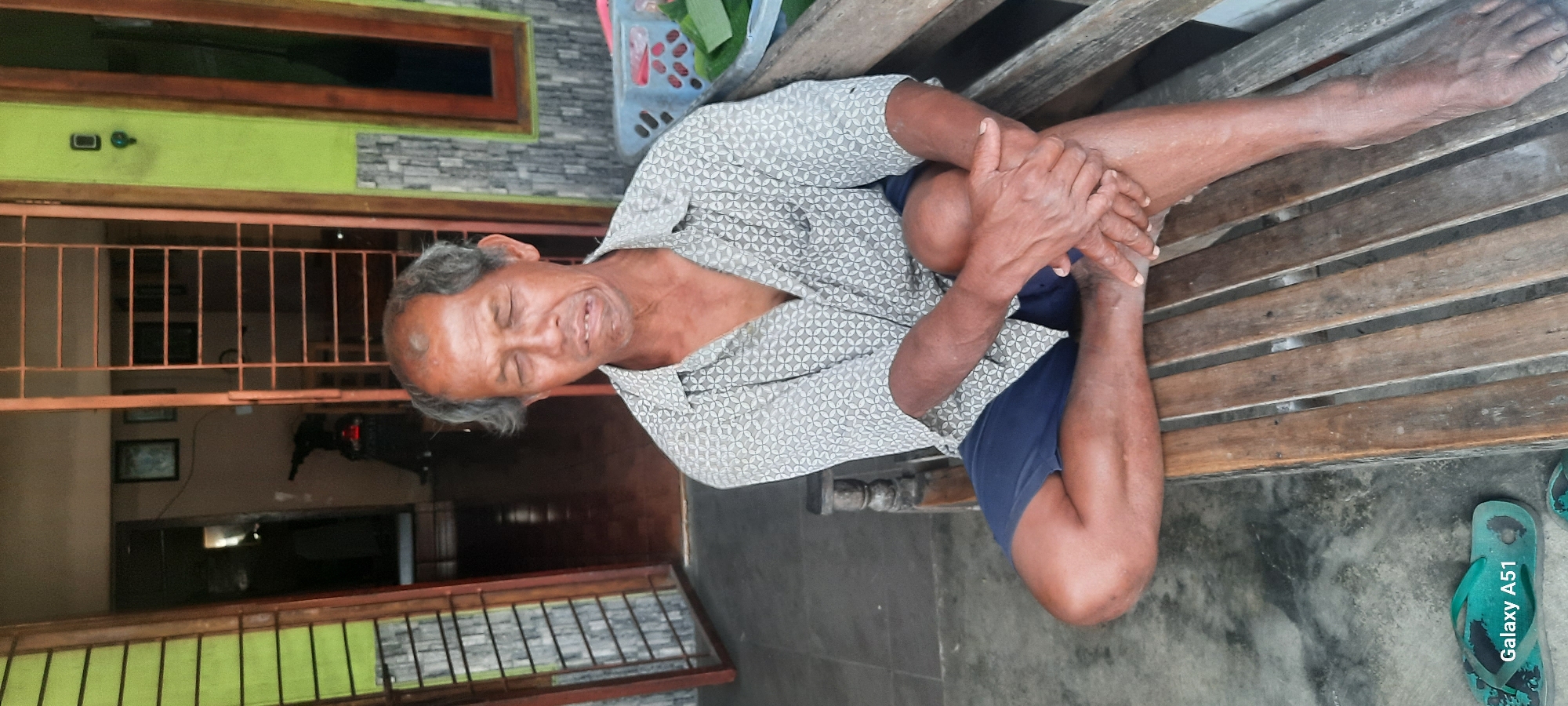 An elderly Indonesian man sits on a weathered wooden bench outside a building. He has graying hair and is wearing a patterned short-sleeved shirt and blue shorts. His eyes are closed, and his mouth is slightly open, with a crooked expression, suggesting he may be experiencing discomfort or pain. His legs are crossed, and his hands are clasped over his knees. The background shows a doorway with a metal security gate, a motorcycle parked inside, and a lime green wall. To the right of the man, a blue plastic basket with green leaves is visible. On the ground to the lower left, a pair of green flip-flops with the inscription "Galaxy A51" is present. The scene appears to be in Selopuro, Indonesia, a rural or semi-rural setting.