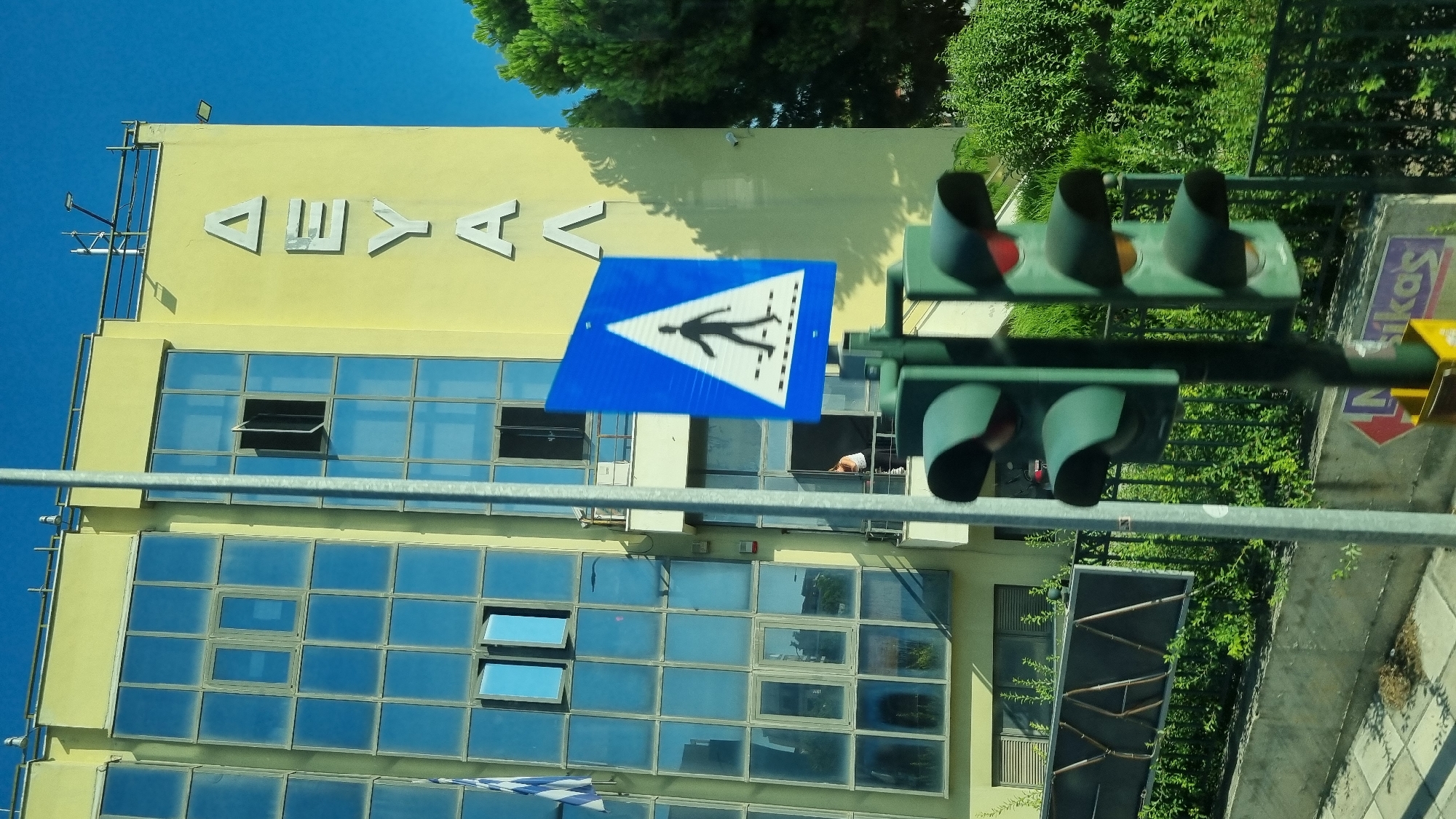 The image shows a building with a light yellow facade and numerous blue-tinted windows. The building has a balcony and a rooftop railing. Greek letters spelling "ΔΕΥΑΛ" are affixed to the facade of the building. A pedestrian crossing sign is visible in the foreground, mounted on a pole. Traffic lights are also present, with the top light illuminated red. A Greek flag is partially visible on the left side of the building. Dense green foliage and trees surround the building. A paved sidewalk is visible at the bottom of the image. The scene suggests an urban environment in Larissa, Greece, possibly in front of a public utility building.