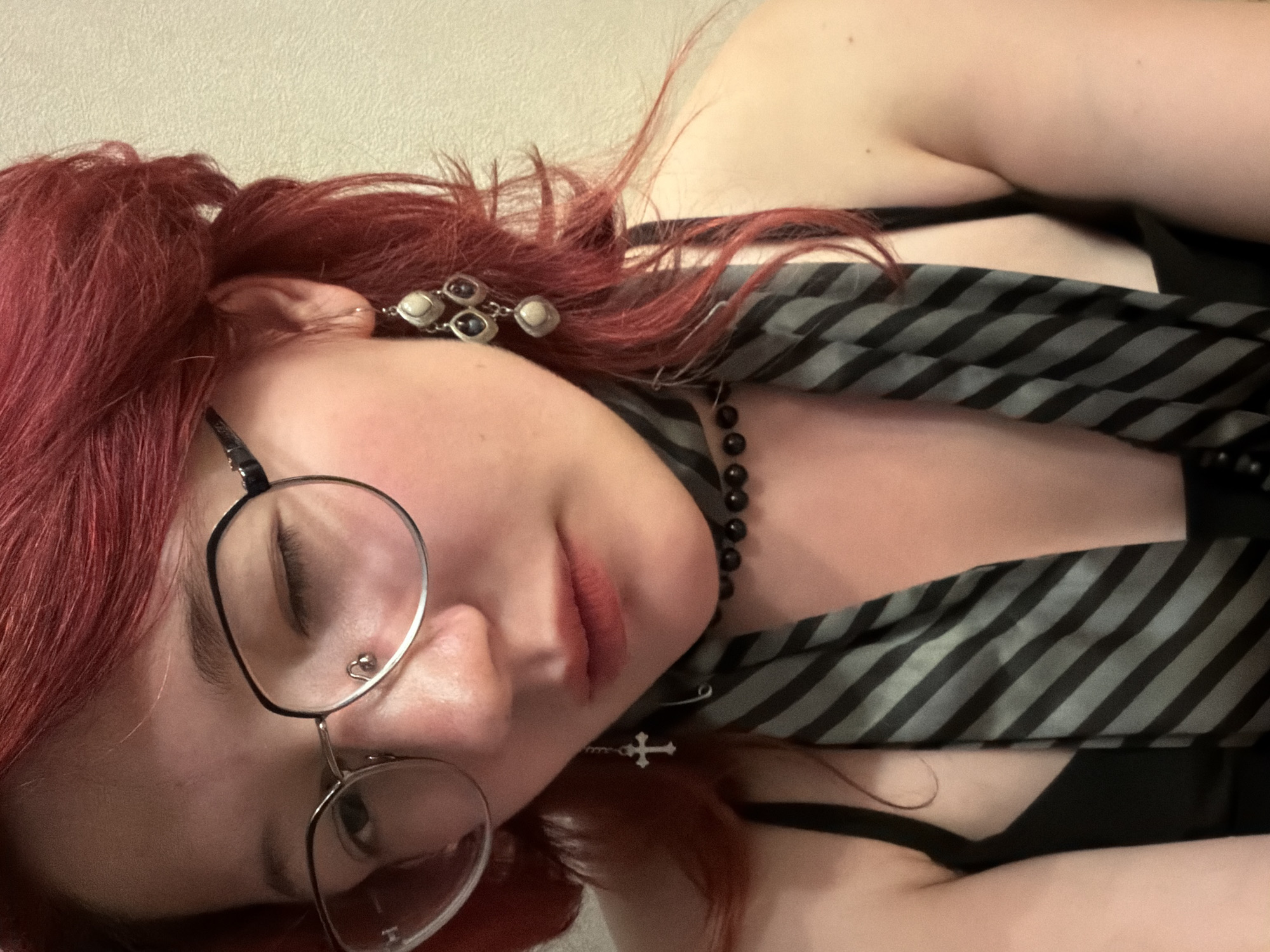 A young person with bright red hair and glasses is looking down, with one eye closed. They are wearing a black top, a grey and black striped tie, a black beaded necklace, and a cross pendant earring. A multi-tiered earring with pearl-like stones is visible on their right ear. The background is a plain, light-colored wall. There is no discernible location within the city of Chelyabinsk, Russia, or any other urban environment.
