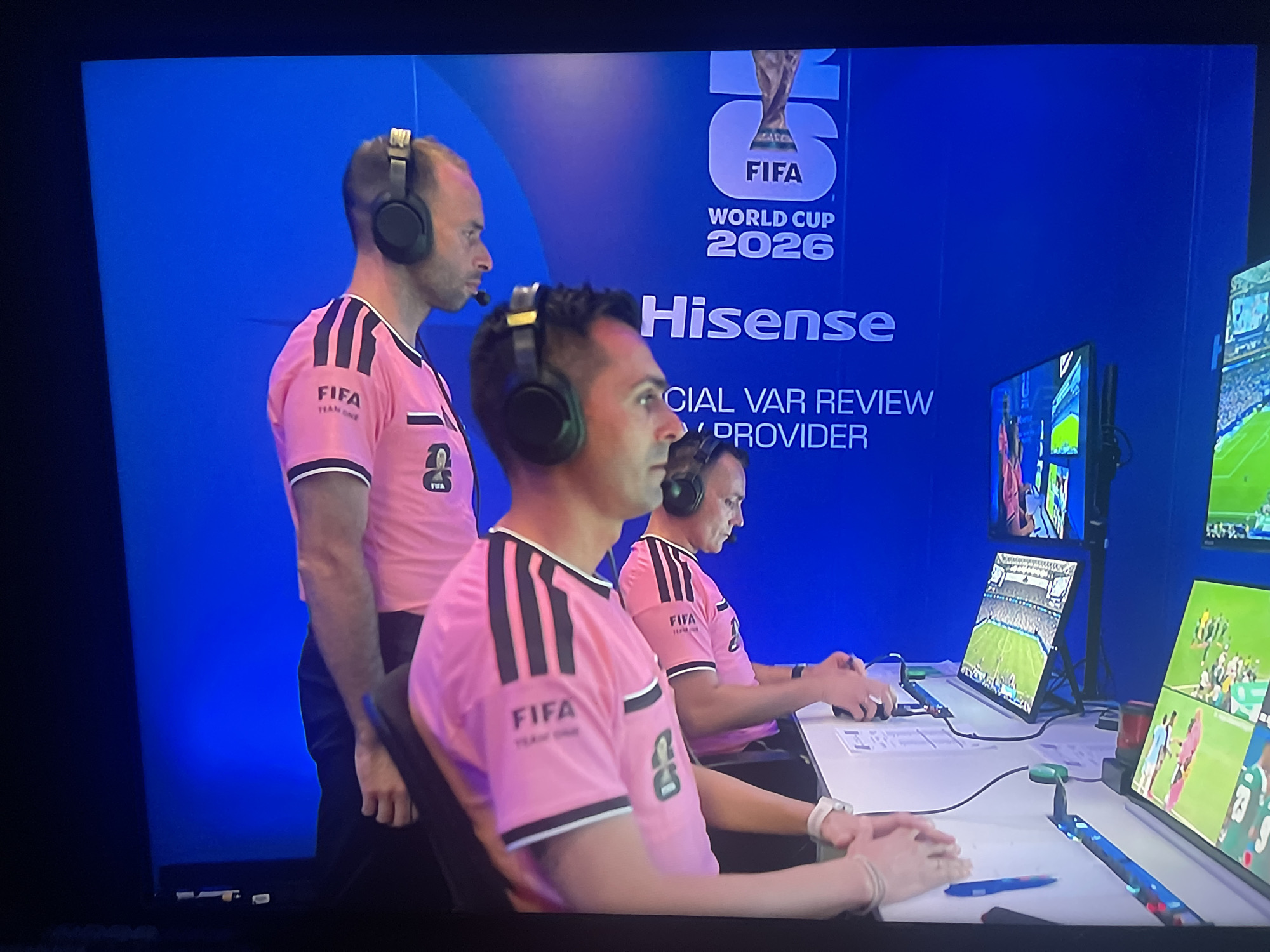 The image depicts a scene likely from a football (soccer) VAR control room, possibly during a training or operational session for the 2026 FIFA World Cup. Three individuals wearing pink FIFA team uniforms with headphones are seated at a table, facing multiple monitors displaying various camera feeds of a football match. The backdrop is a bright blue wall with the "FIFA World Cup 2026" logo and the "Hisense" brand prominently displayed, indicating a sponsorship. The individuals appear to be focused on the screens, with one person's hands on a keyboard or control device. The environment suggests a professional setting for sports officiating technology. This specific location is identified as Ruse, Bulgaria.