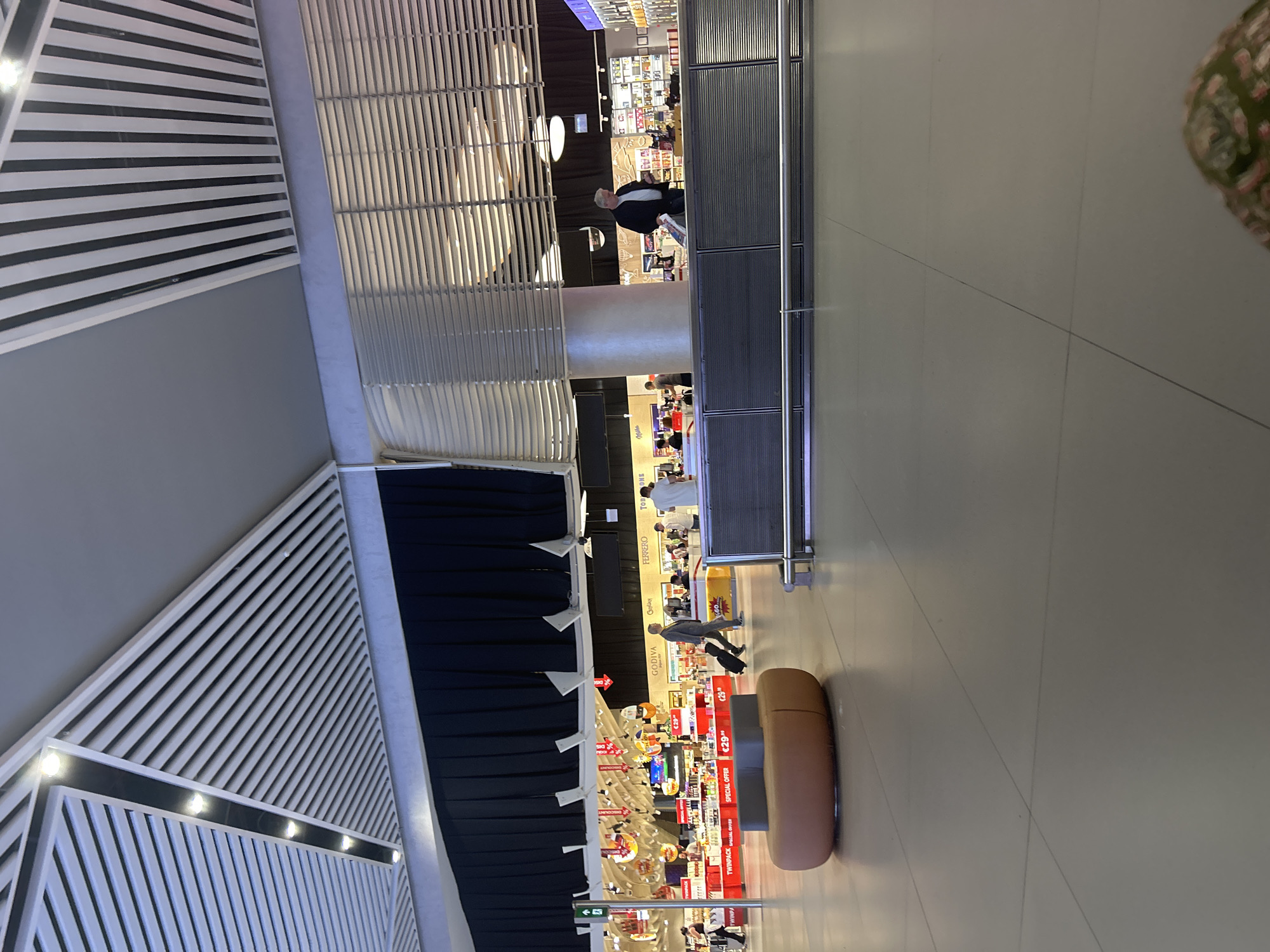 The image depicts an interior scene, likely an airport terminal in Arnavutköy, Turkey, characterized by modern architecture. The ceiling features a distinctive design with alternating white and dark strips, some of which are illuminated by embedded lights. Below, a large space with a tiled floor suggests a public area. To the left, a black fabric curtain hangs, partially obscuring shops or displays behind it. A man in a suit is seen walking with a rolling suitcase, indicating travel. In the mid-ground, a counter area displays various products, with several individuals interacting or browsing. An orange, oval-shaped bench is positioned in the foreground, adding to the seating arrangements within the terminal. The overall impression is one of a bustling, well-lit commercial space designed for travelers.