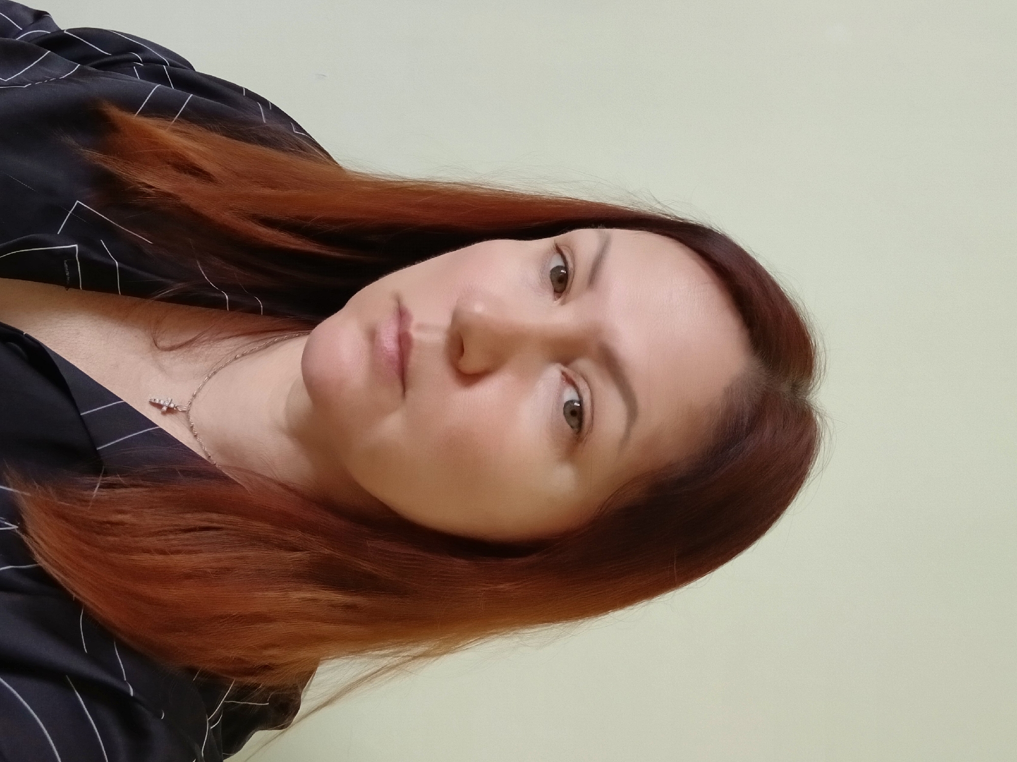 A selfie of a woman with auburn hair is depicted indoors against a light green background. She is wearing a black top with white stripes and a silver necklace with a cross pendant. Her gaze is directed towards the camera. The location is Unknown, Unknown.