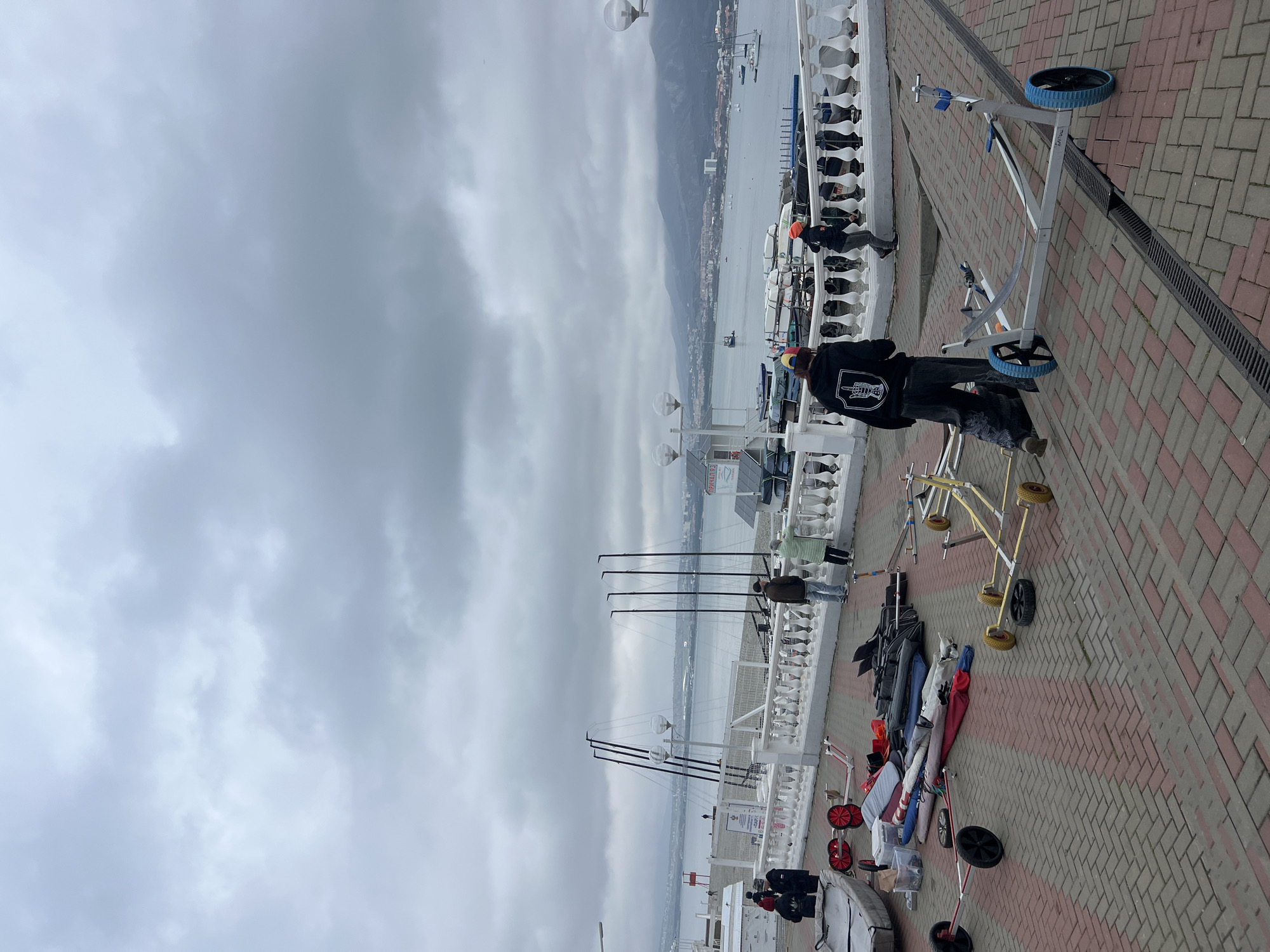 The image depicts a waterfront scene in Gelendzhik, Russia, specifically along a promenade or pier area overlooking Gelendzhik Bay. The sky is overcast with heavy grey clouds, and the bay's water appears calm and grey-blue.
In the immediate foreground, a paved area with red and brown interlocking pavers contains a collection of sailing equipment. This includes a grey dinghy or small boat, several boat launching trolleys (one grey with red wheels, one yellow with small yellow wheels, and another grey with larger blue wheels), rolled sails, black equipment bags, and various spars and boat parts laid directly on the ground. Drainage grates are embedded in the pavement.
Several individuals are present. One person, seen from behind, wears a dark hoodie with a white hand gesture logo on the back (depicting a middle finger) and dark pants, walking towards the right side of the frame near a grey launching trolley. To their right, another person in an orange cap is partially visible, bending near a white balustrade. Further left, two individuals, including a child, are near the grey dinghy. Towards the center-right, two adults, one in a brown jacket and the other in a light green jacket, stand at a white balustrade, looking out over the water. Other indistinct figures are visible further down the promenade and near the water's edge.
Beyond the paved area, a white balustrade with spherical lamp posts separates the promenade from a lower beach or storage area. This area features several tall, dark masts standing upright and numerous small boats stored near the water. A small white building with a blue roof and the text "ПРИЧАЛ 93" (PIER 93) is visible to the right.
The Gelendzhik Bay is visible in the mid-ground, dotted with various boats, including sailboats and motorboats. The distant background features a cityscape along the shoreline, backed by large, forested mountains partially obscured by the low-hanging clouds.
