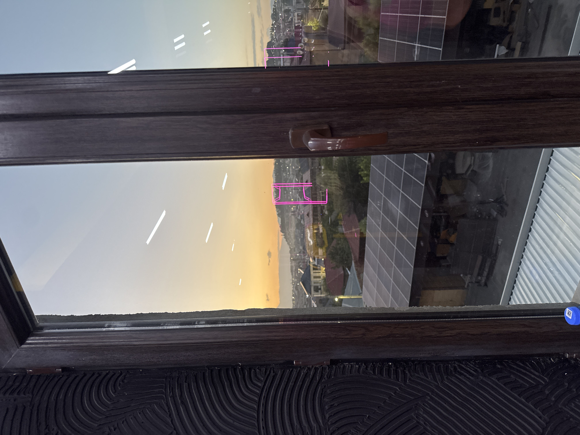 The image shows a view through a window overlooking the city of Arinj, Armenia, at sunset. The sky is a gradient of orange and blue, with the sun low on the horizon. In the foreground, a dark, textured wall is visible to the left, and a wooden window frame with a brown handle is in the center. Reflected in the glass are pink geometric shapes and horizontal white lines.
The cityscape below features a variety of buildings with red and brown roofs. Solar panels are visible on the rooftops of some structures. In the distance, hills and mountains form the backdrop. The overall impression is a tranquil evening scene in a residential area.