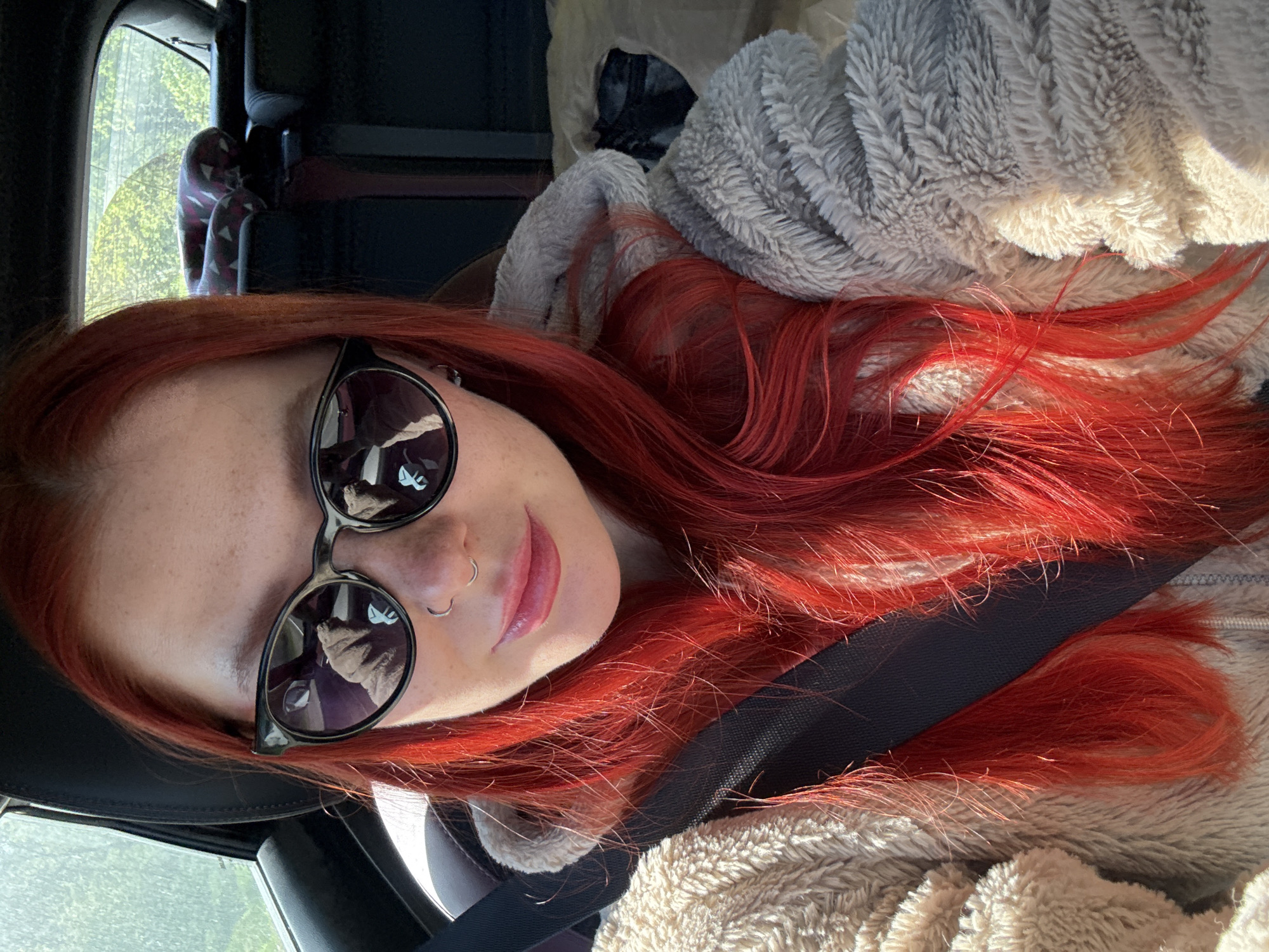 A selfie taken by a woman with red hair and sunglasses, likely inside a car. The woman is wearing a seatbelt and a fuzzy gray jacket. The location is not identifiable as Pokrovskoye, Russia from the given visual information.