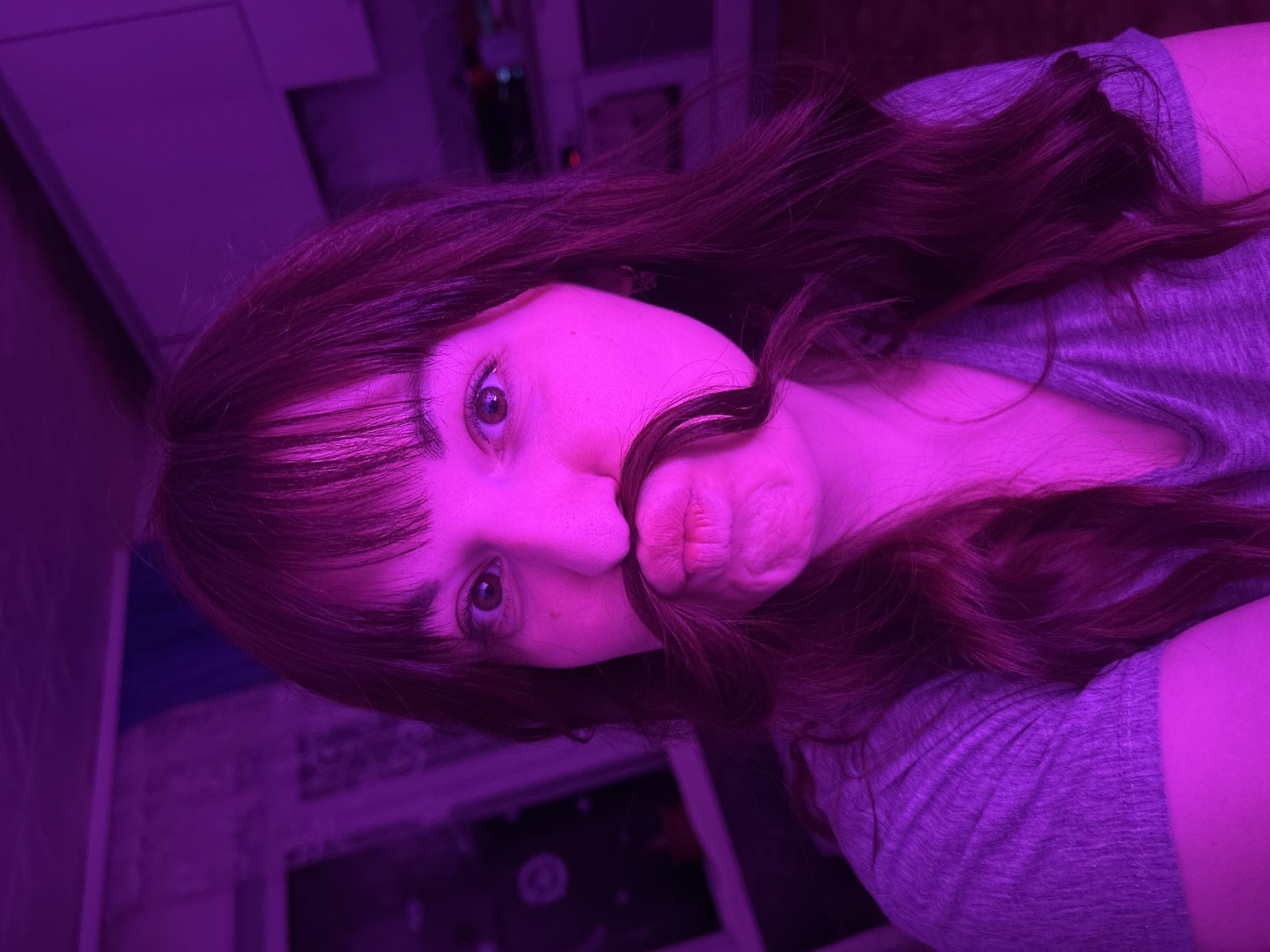 A woman with brown hair, bangs, and fair skin is shown from the chest up. She is lying on her side, facing the camera. Part of her hair is pulled to her upper lip to mimic a mustache. She wears a gray t-shirt. The lighting is purple. A window and wall are in the background. The location is not discernible.