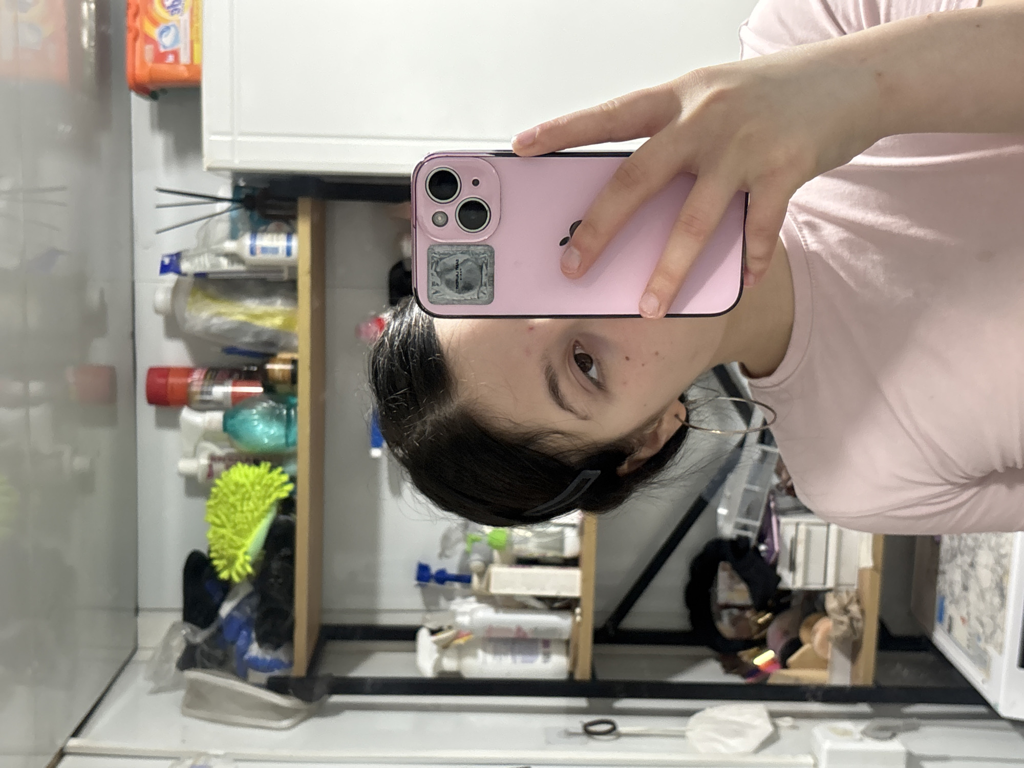 A woman with dark hair and a pink t-shirt is taking a selfie in a bathroom. She is holding a pink smartphone, obscuring part of her face. The phone has a clear sticker on the back. Behind her are shelves containing various household cleaning products and toiletries. A white cabinet is visible above the shelves. Other objects in the background include a hair clip in her hair, a hoop earring, a vanity with makeup, and a black circular object. The specific location in Фоки, Russia, cannot be determined from the visual information provided.