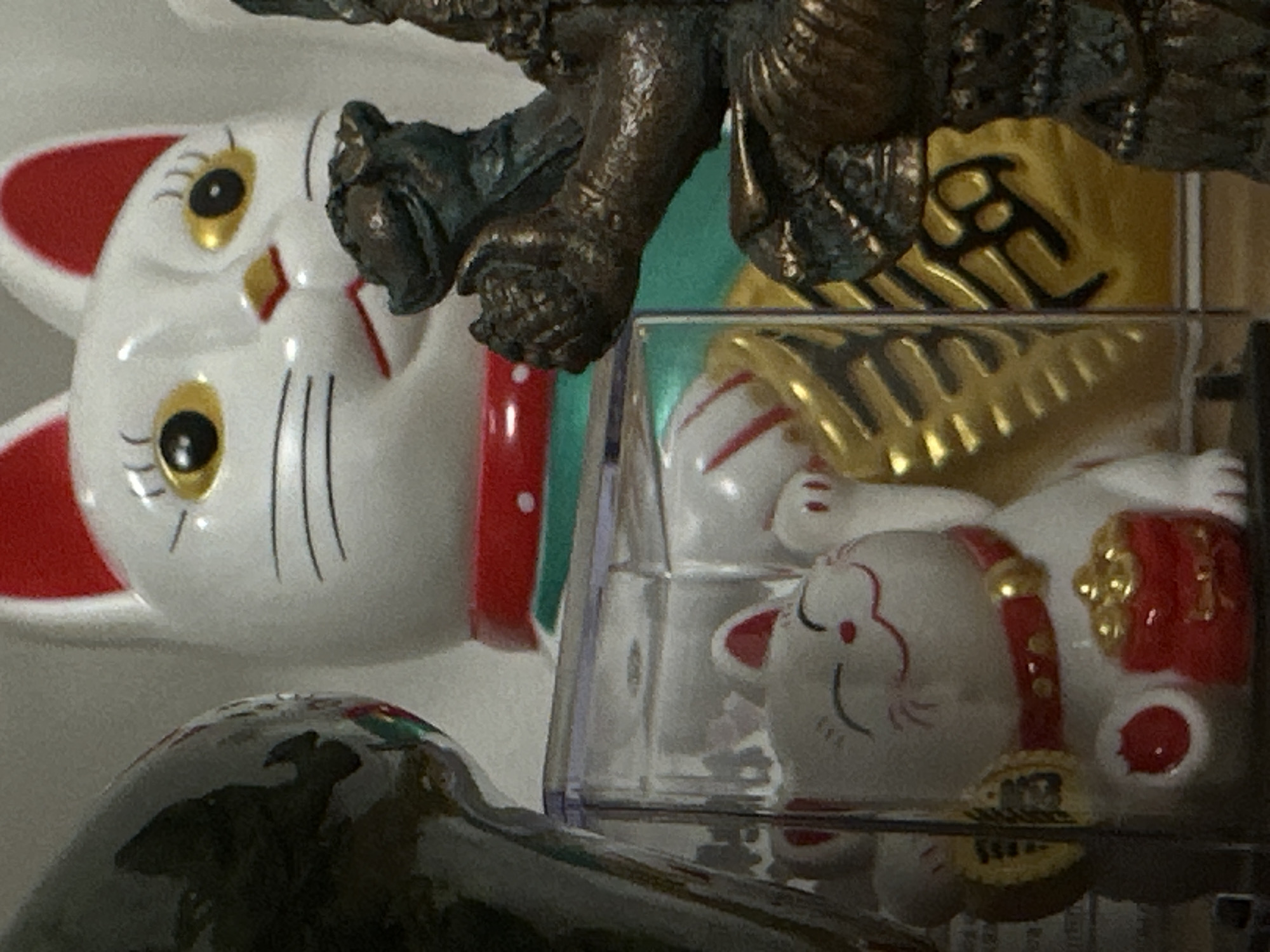 The image features three lucky cat figurines. Two are visible on the left, one with a white body, red ears, and golden eyes, and the other is flipped sideways with closed eyes. A third cat figurine, also white with red and gold accents, is enclosed in a clear plastic box. Above the figurines are parts of a statue with a dark metallic finish. The background is blurred, but the setting appears indoors. There is no indication that the picture was taken in Rostov-on-Don, Russia, and no information about people or their actions is visible in the photo.