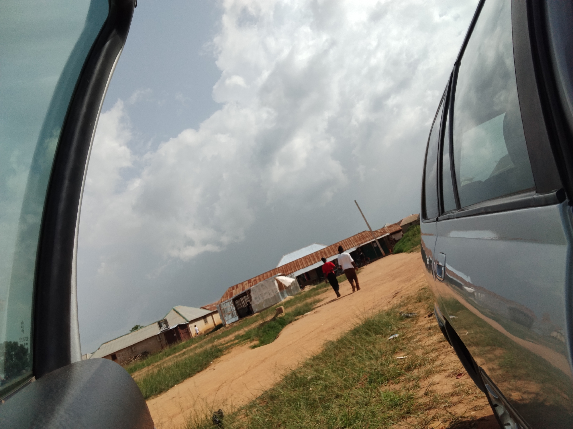 The image captures a scene in Durumi, Nigeria, with a view from inside a vehicle looking out at a dusty road and several buildings. The sky is overcast with dramatic clouds. On the road, two individuals are walking away from the camera. One person is wearing a red shirt and dark pants, while the other is in a white shirt and brown pants. To the right of the road, there is a light-colored vehicle, possibly a van, with its side facing the viewer, reflecting the cloudy sky and the surroundings. The buildings in the background appear to be simple structures with corrugated metal roofs. The environment suggests a rural or semi-urban setting. Grass and dry vegetation line the sides of the road.