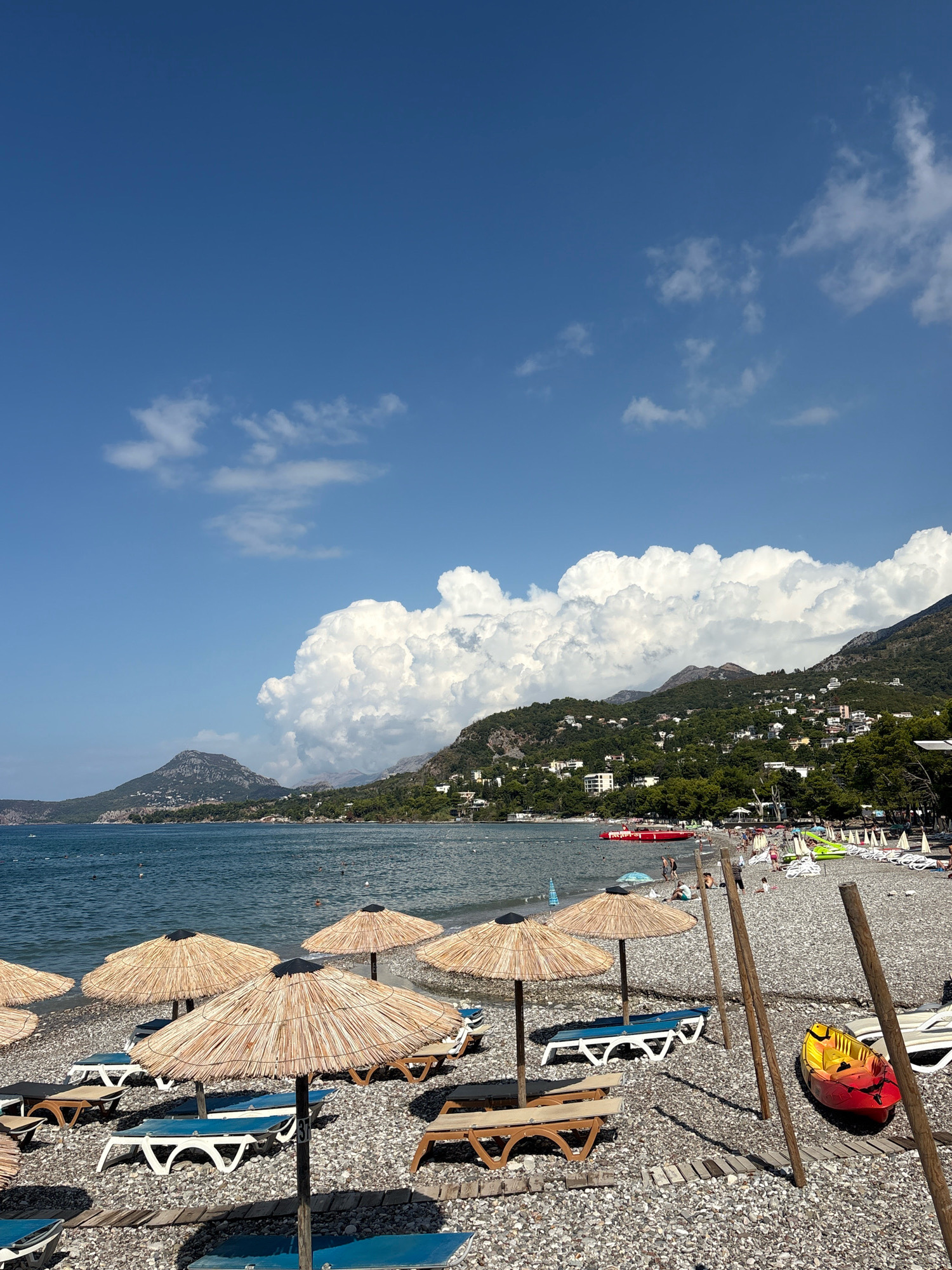 This is a photograph of a pebble beach in Bar, Montenegro, on a sunny day with a partly cloudy sky. The foreground features rows of unoccupied beach chairs and thatched umbrellas. A yellow and red kayak rests on the pebbles near the center-right of the image. In the background, the calm blue sea stretches towards a mountainous coastline covered in dense green vegetation and dotted with white buildings. The sea has some people visible in the water. The sky is a vibrant blue with large, fluffy white clouds dominating the upper right portion of the frame.