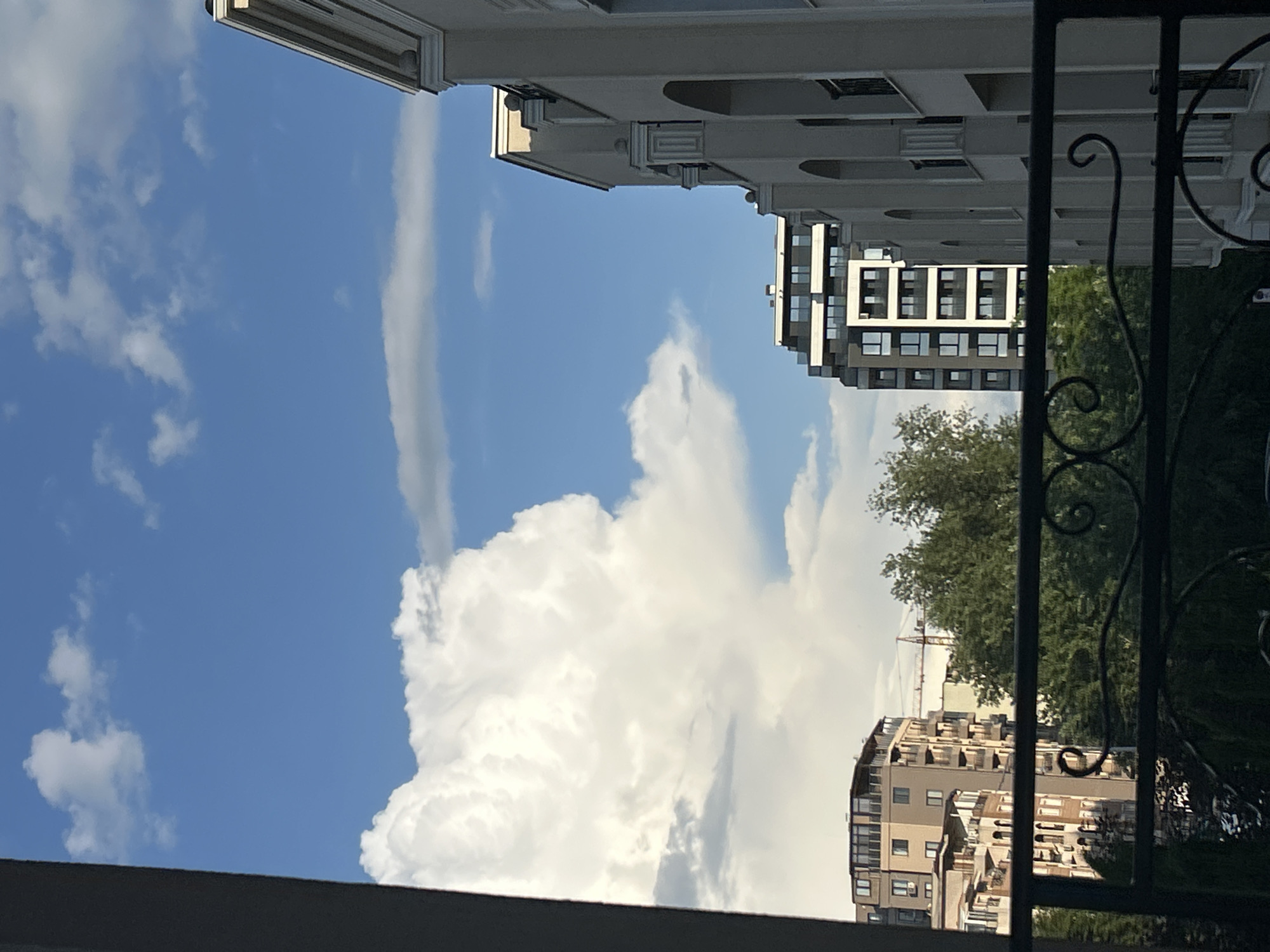 The image, taken from a low-angle perspective, reveals a view of the sky and nearby buildings in Plovdiv, Bulgaria. The sky is predominantly blue, filled with white cumulus clouds of varying sizes and shapes. Two buildings are visible: one with a modern architectural style, featuring a grid-like facade, and another with a more traditional design and ornate architectural details, including archways. The buildings are positioned as if facing each other. In the distance, a construction crane can be seen over the treeline. A portion of a wrought-iron fence is visible in the right part of the frame, slightly out of focus. The lower section of the image has a horizontal plane (likely part of the structure from which the picture was taken).