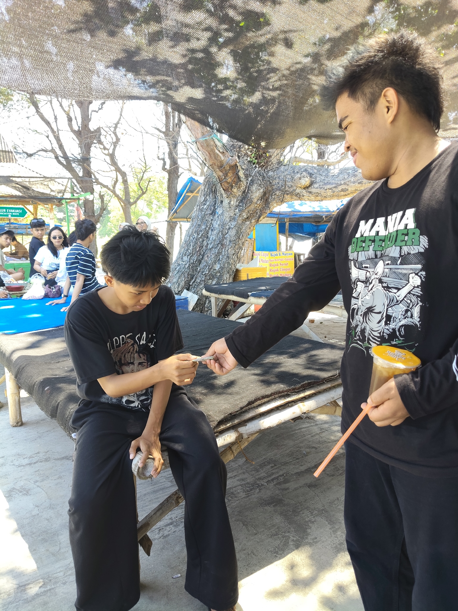 Two young men are outdoors in what appears to be a beachside location in Camplong, Indonesia. The man on the right, wearing a black long-sleeved shirt with a graphic print, is holding a plastic cup with a straw and offering a cigarette to the man on the left. The man on the left, dressed in a black t-shirt and black pants, is seated on a low, rustic bench covered with a dark fabric. He is looking down and has a cigarette in his hand, seemingly accepting it. The background shows trees with a rough canopy, a beach or sandy area, and some stalls or structures that suggest a public space, possibly a recreational area. The lighting indicates it is daytime, likely sunny. Other people are visible in the background, some seated and others standing, indicating a social gathering.