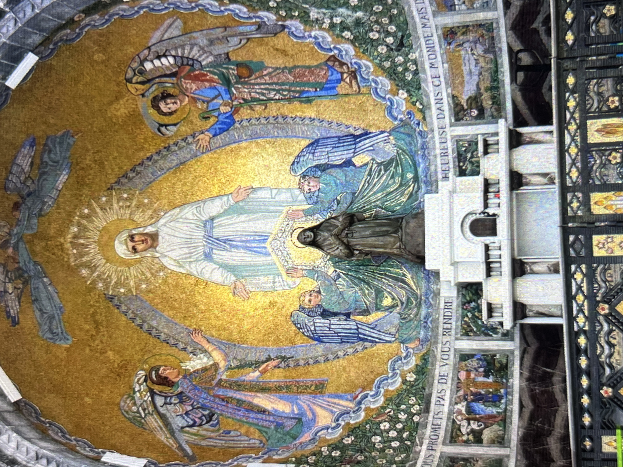 The image depicts a mosaic of the Virgin Mary ascending to heaven, surrounded by angels, likely located inside the Santuario de la Divina Pastora in Tinaquillo, Venezuela. The mosaic is predominantly gold, featuring Mary in a white robe with a blue sash. Angels in blue and green garments are positioned around her. Above and below Mary, angels bear long, serrated objects that arc around the central figures. The mosaic is bordered by text in French and is on the interior surface of a structure. Additional mosaics and the interior architecture of the sanctuary frame the central depiction.