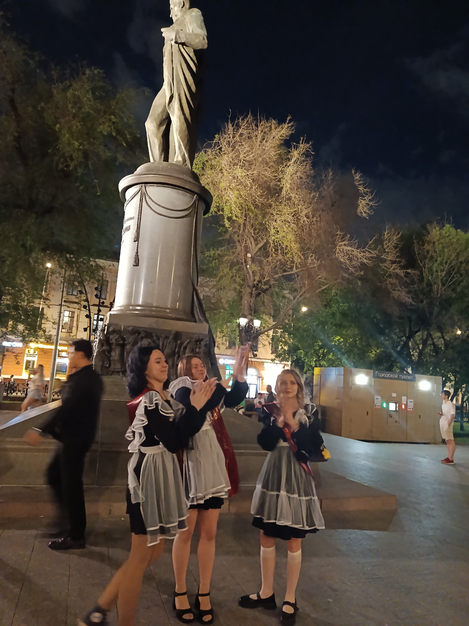 The image captures a night scene in an urban environment. In the foreground, three young women dressed in what appear to be school uniforms stand on a stone platform, and appear to be clapping. They wear dark dresses with white aprons and red sashes. Behind them, mounted atop the platform, is a statue of a man draped in fabric. In the background, the scene includes buildings, trees, streetlights, and other pedestrians. A sign that reads “Городской туалет” (City Toilet) is visible.