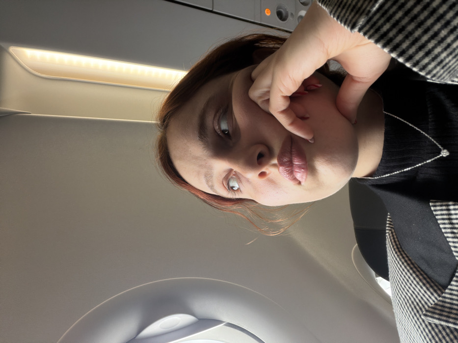 The image displays a female individual inside an airplane cabin. The scene is provided with the contextual location of Tuzla, Turkey. The subject, with reddish-brown hair, is positioned in the foreground, facing slightly right but looking upwards and to the left. Her left hand is resting with fingers lightly curled against her chin/jawline. She wears a black turtleneck or similar top, a black and white checkered blazer, and a silver-colored chain necklace with a small, light-colored pendant. Her fingernails are painted light red.
The immediate environment is an aircraft interior, specifically a window seat. A circular airplane window is visible on the upper left, framed by the light-colored interior paneling. The exterior view from the window is not discernible. The light-colored ceiling and overhead paneling are prominent. An illuminated strip of LED-like lights runs along the overhead panel on the right side of the image, casting a warm glow. An orange indicator light is visible on an overhead control panel on the far right. A dark headrest of an airplane seat is partially visible behind the person's right shoulder. No other individuals are visible, and there are no explicit interactions with other people.