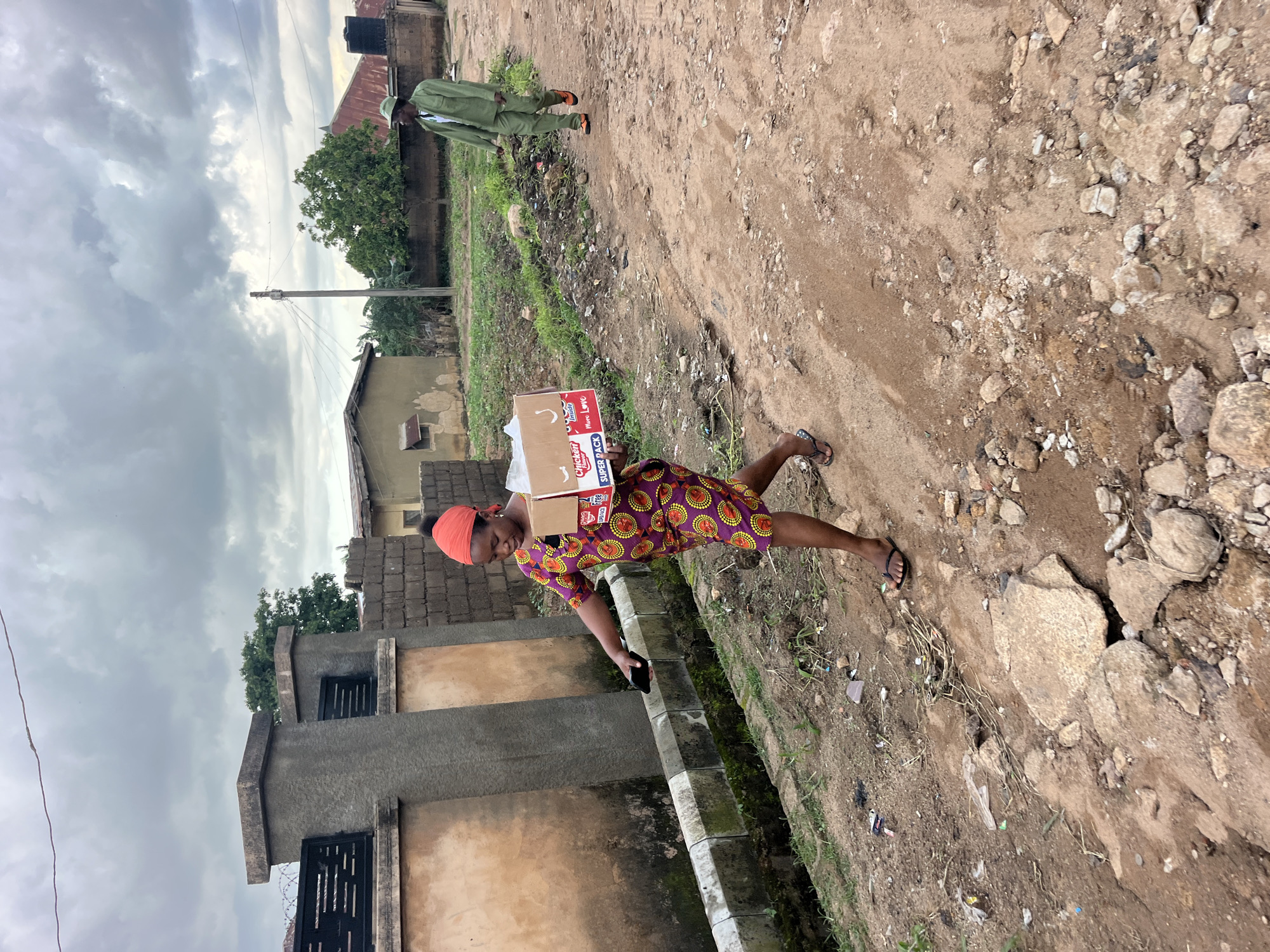 A woman walks on a dirt path carrying a cardboard box on her back. She is wearing a red headwrap and a colorful patterned dress. In front of her, a person in a green uniform walks away from the camera. The ground is rough and rocky with patches of grass and debris. Buildings and trees are visible in the background. The sky is overcast and cloudy. The scene appears to be a rural or semi-urban environment. The text on the box reads "Chicken SUPER PACK More Love". The overall impression is that of a delivery or transport of goods in an outdoor setting. The time of day is difficult to determine, but the overcast sky suggests it is likely daytime.