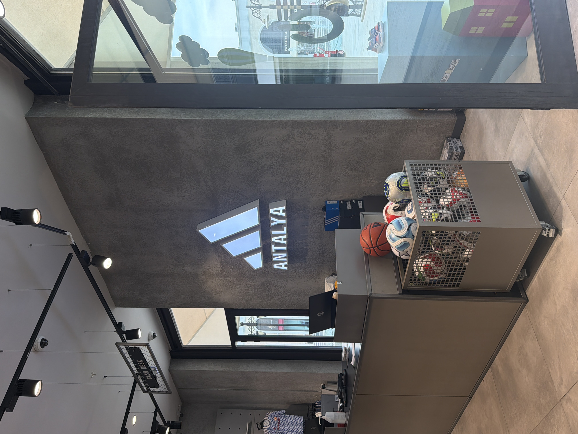 The image shows the interior of an Adidas store, likely in Serik, Turkey, as indicated by the "ANTALYA" text on the wall, which is a region that includes Serik. The store features a grey textured wall with a prominent illuminated Adidas logo and the word "ANTALYA" below it. In the foreground, there is a checkout counter with laptops and a hand sanitizer dispenser. Next to the counter, a metal cart on wheels is filled with various sports balls, including soccer balls and a basketball. To the right, a large glass window displays shoes on a pedestal and a colorful model house. The overall environment is clean, modern, and well-lit, typical of a retail sports store. There are no people visible in the immediate foreground or at the counter. The background shows a glimpse of an outdoor shopping area through the store's entrance.