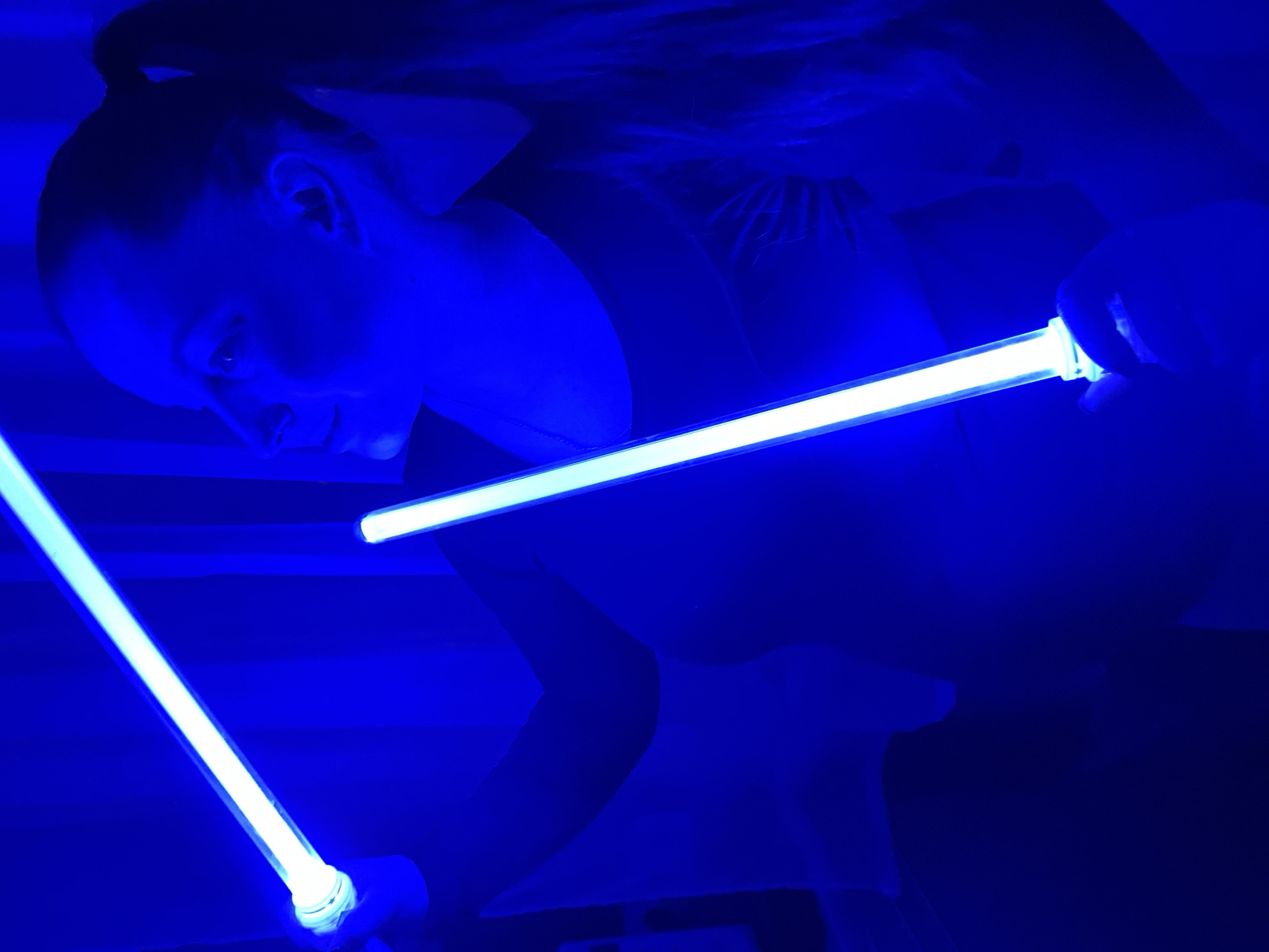 A woman with her hair pulled back is shown in the image. She is holding two blue light sticks, one in each hand. The scene is illuminated by the blue light, giving the overall image a monochromatic look. It is difficult to determine the exact location from the image, and there is no information to suggest this scene is located in Izhevsk, Russia.