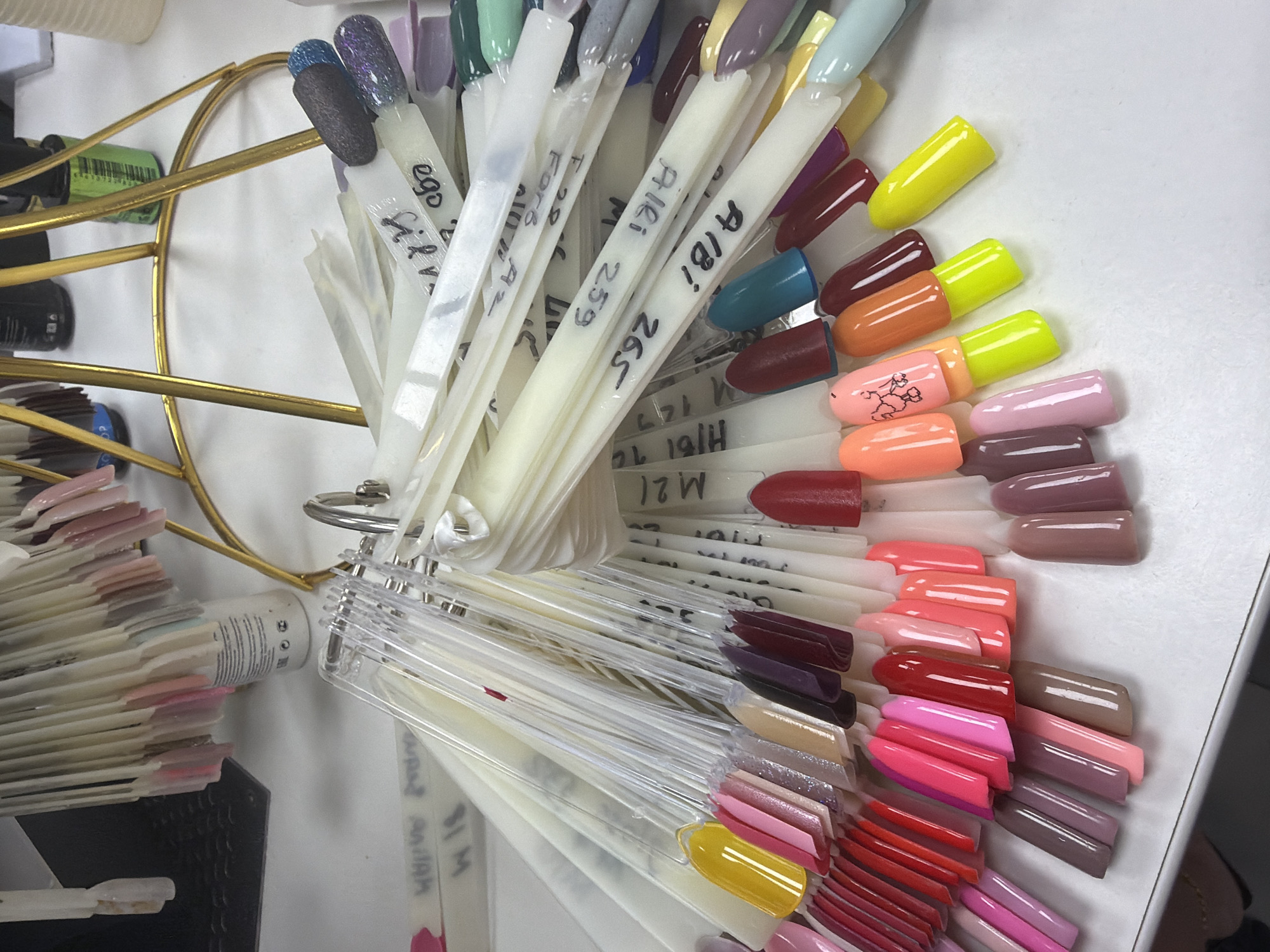 The image displays a close-up, eye-level, indoor shot of a table filled with nail polish swatches. The swatches are arranged on fan-like displays, with many individual plastic sticks holding different colored nail polish tips. Some of the sticks have handwritten labels, such as "A181 265," "A181 259," and "ego." Various colors are visible, including reds, pinks, purples, browns, yellows, oranges, blues, and greens. In the background, a gold metal stand holds more swatches, and several bottles, one with a green label and another with a blue label, are visible on the stand. The overall environment appears to be a nail salon. The scene is static, with no visible action or interaction. The location is Budva, Montenegro.