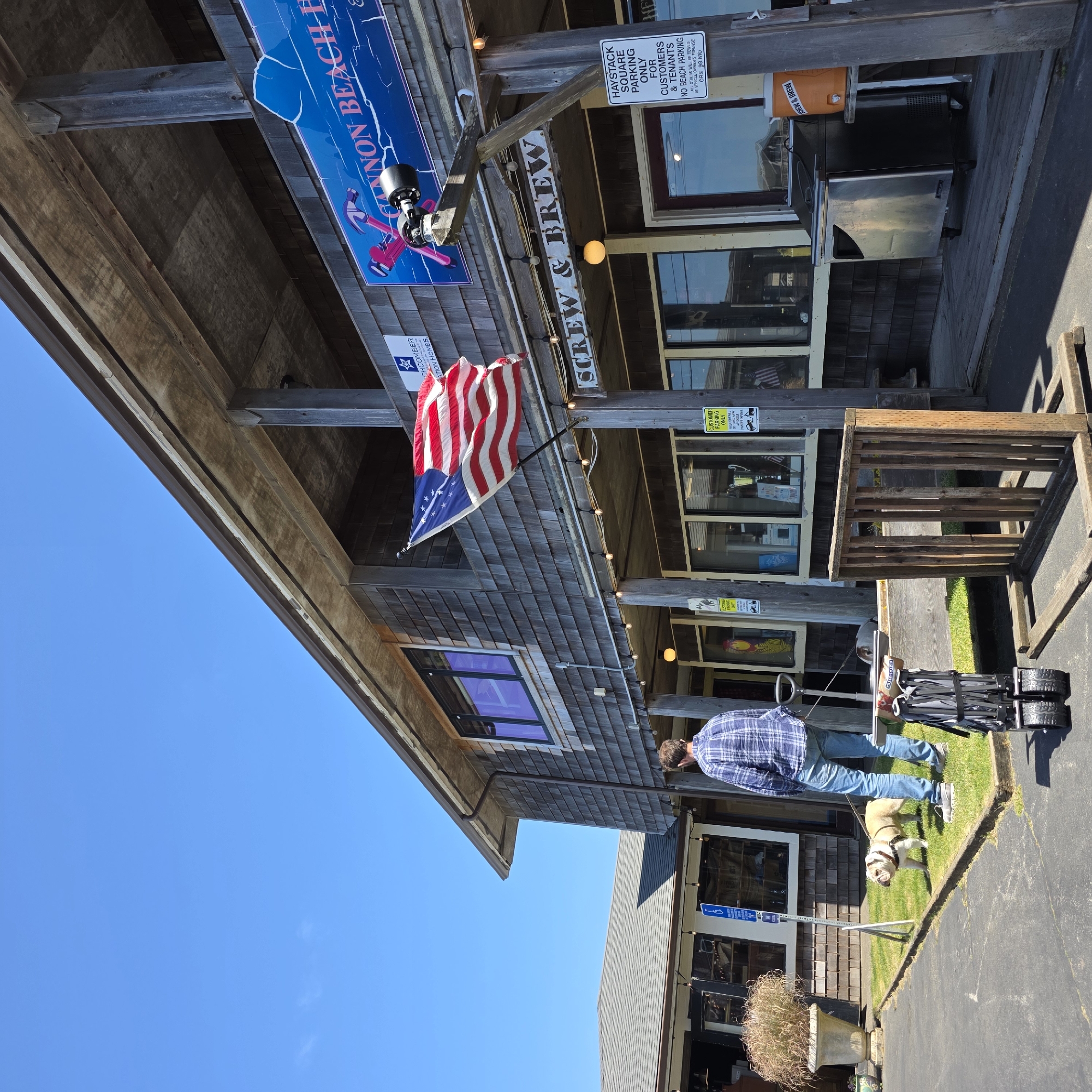 An American flag flies on a pole outside a wooden building with shingled siding. A person wearing a blue plaid shirt and jeans stands on a grassy area, holding a leash attached to a bulldog. Next to the person is a folded collapsible wagon. A sign on the building reads "SCREW & BREW." Another sign indicates "HAYSTACK SQUARE PARKING ONLY FOR CUSTOMERS & TENANTS." An orange water cooler with a "BREW & BREN" sticker sits on a table. The scene is set against a clear blue sky on a sunny day.
