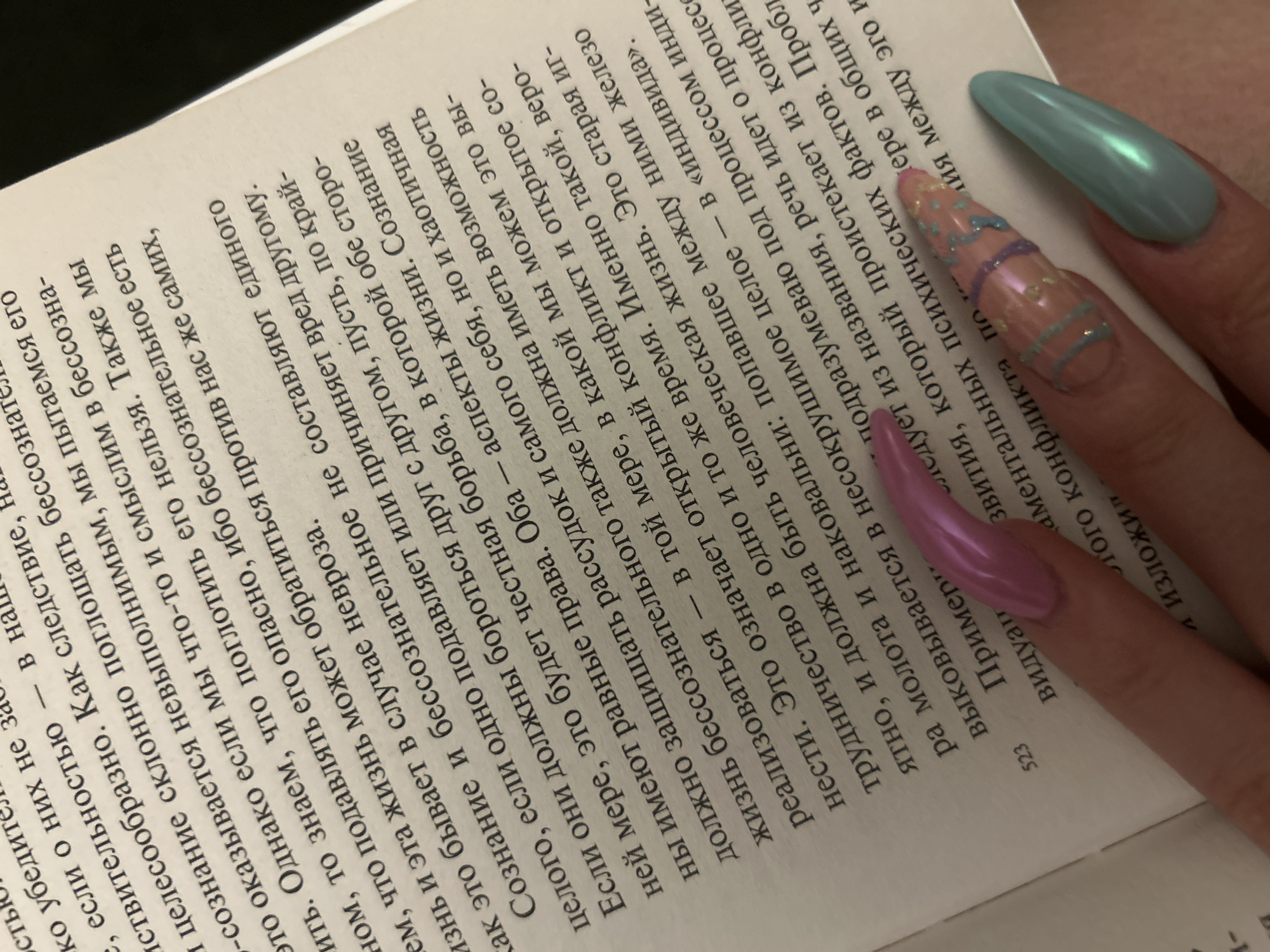 Here is a detailed description of the image:
The image shows an open book with text in Russian. The book's pages are off-white, and the text is printed in black. The left-hand page is visible, with a partial view of the right-hand page. The text appears to be a discussion of psychological concepts, with words like "сознание" (consciousness), "бессознательное" (unconscious), and "психических" (psychological) visible.
In the foreground, three manicured nails are prominently displayed. They are long and have elaborate designs. One nail is a solid light blue, another is a pale pinkish tone with pastel blue and purple stripes, and the third nail is a lavender shade. The nails are held over the book, partially obscuring the text. The book's page number "523" is visible.
The image is tightly cropped, focusing on the book and the nails, and the angle is slightly tilted. The overall impression is of someone reading a book with carefully styled nails.