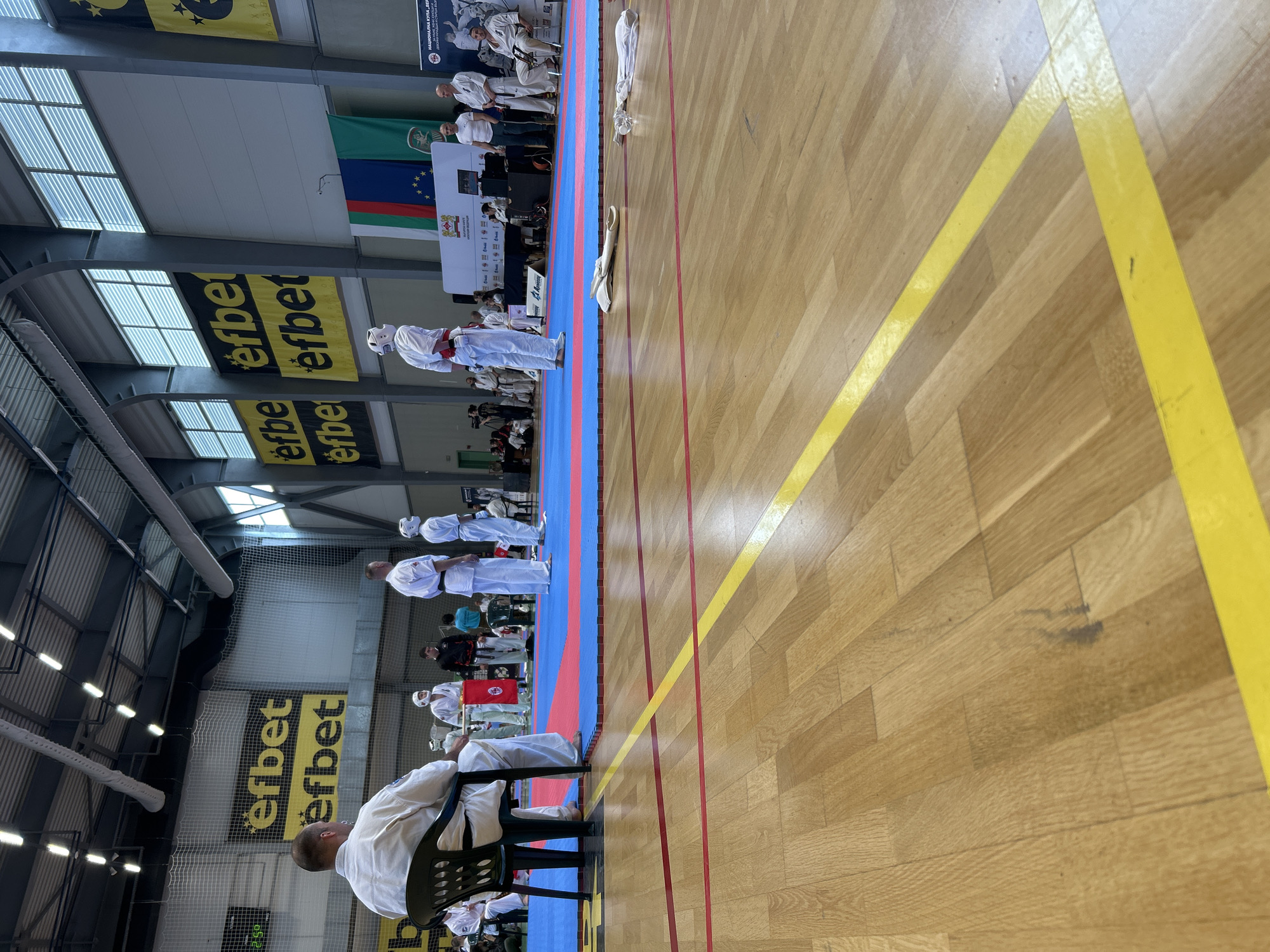 The image shows an indoor martial arts event in Stara Zagora, Bulgaria. Several individuals in white martial arts uniforms are lined up on a blue and red mat. One person is seated on a black plastic chair, also wearing a white uniform. Banners with "efbet" are visible in the background, along with the flags of Bulgaria and the European Union. The flooring is wooden with yellow and red lane markings. The scene is a martial arts competition or demonstration taking place inside a large hall with a high ceiling and skylights.