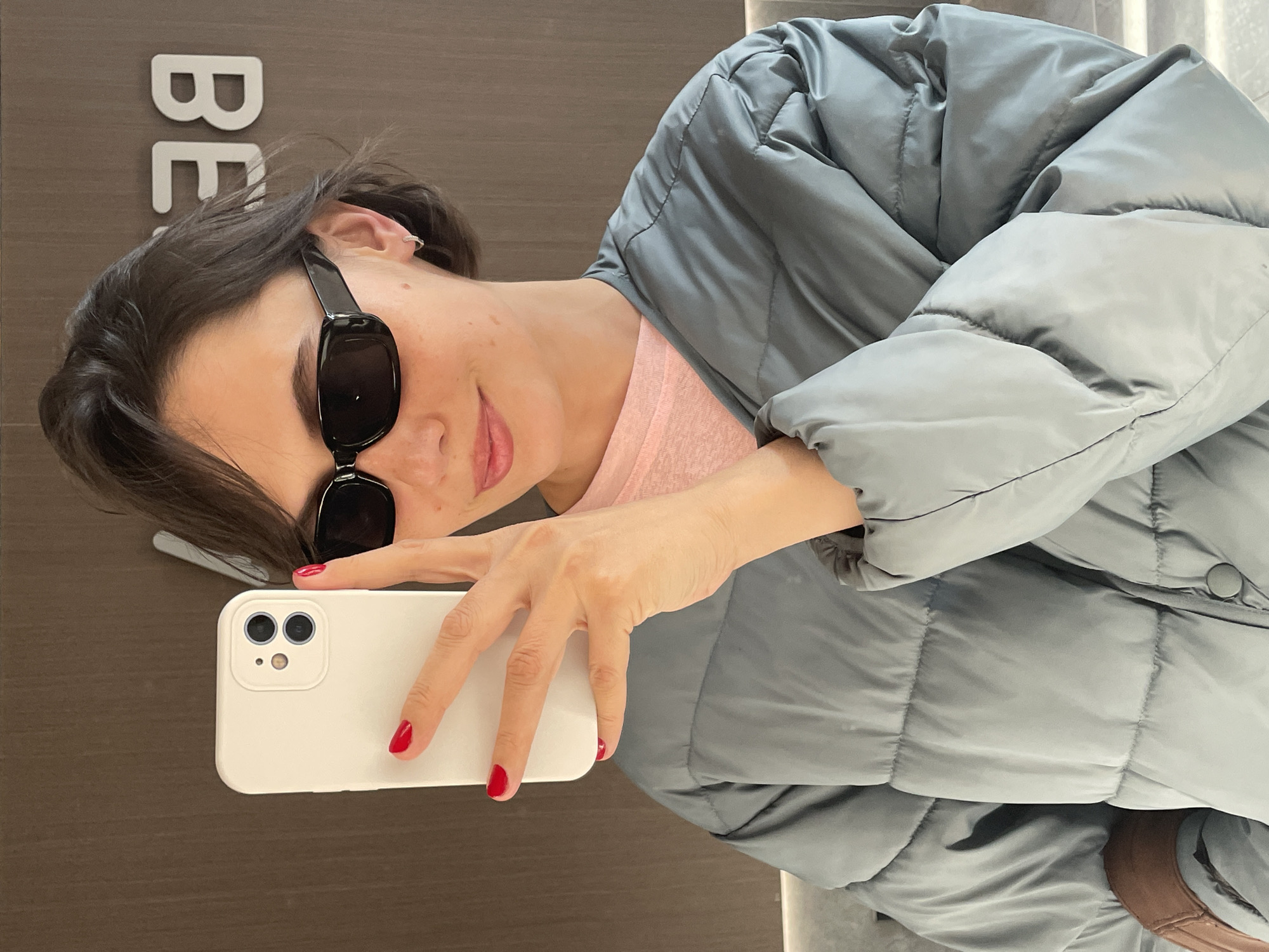 A woman wearing sunglasses and a light blue puffer jacket is taking a selfie with a white smartphone. She is indoors. The letters "BE" are visible in the background, which is a wood-paneled wall. The woman has red nail polish. The location appears to be in Izhevsk, Russia.