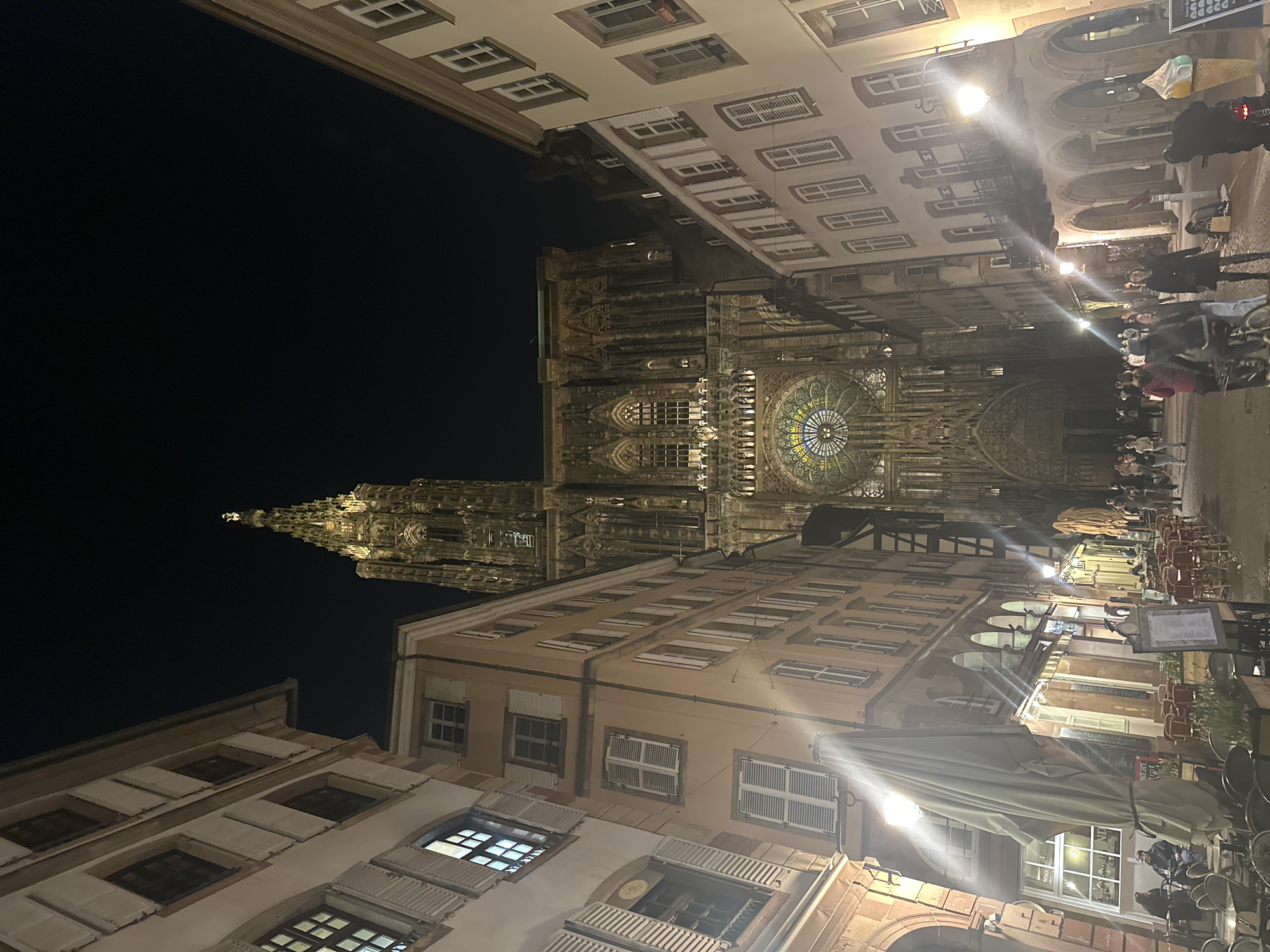The image depicts a night scene in Strasbourg, France, featuring the Strasbourg Cathedral prominently. The camera angle looks upward, showcasing the cathedral's intricate architecture against a dark sky. Buildings line both sides of the street, illuminated by streetlights that reflect off the wet pavement. People are present along the right side of the image, walking and interacting in the pedestrian area. Tables and chairs from nearby cafes occupy the sidewalk, suggesting a vibrant urban environment.
