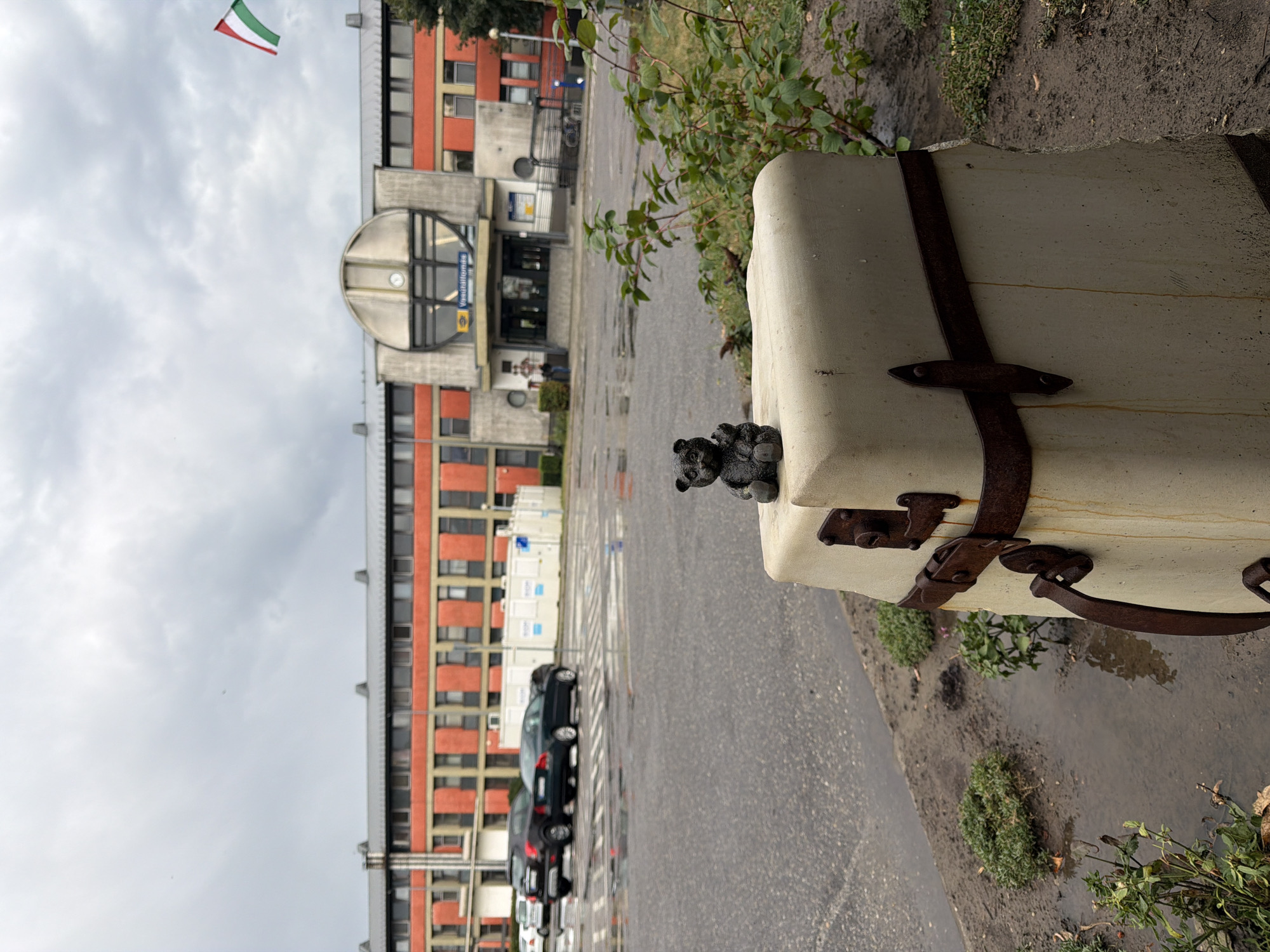 This is a photograph taken at the Záhony train station in Záhony, Hungary. The image is captured from a low angle, focusing on a small, dark-colored bear figurine seated on a light-colored, stone-like pedestal that resembles a suitcase. The pedestal has two rusty metal straps around it, one near the top with a latch mechanism and another near the bottom with a curved handle. To the right of the pedestal, green foliage from a bush is visible.
In the background, the train station building is prominent, characterized by its red brick facade and a large, circular, metallic structure above the entrance. A sign above the entrance reads "Vasútállomás" (meaning "Train Station" in Hungarian). A Hungarian flag, red, white, and green, flies from a pole to the right of the building.
The foreground shows a wet, asphalt-covered area, indicating it might have recently rained or is raining, with several puddles reflecting the overcast sky. Several cars are parked or driving in the mid-ground, and in the far distance, more of the train station structure and trees are visible. The sky is heavily clouded and grey. No people are clearly depicted, though a lone figure can be seen standing near the entrance of the station in the background. The overall scene suggests a somewhat gloomy, damp day at a functional train station.