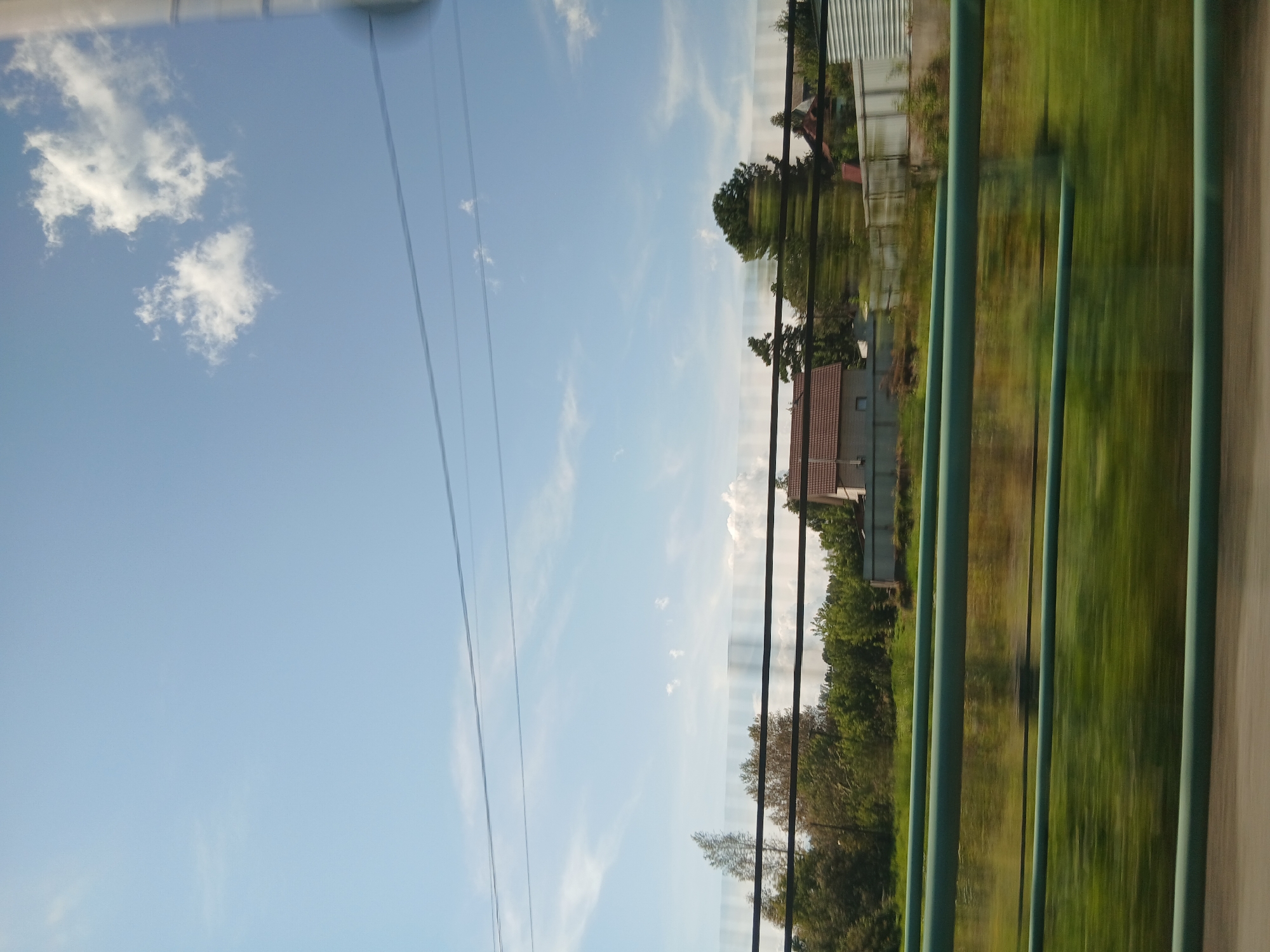 The image depicts a blurry, motion-filled view of a rural or suburban landscape under a bright blue sky with scattered white clouds. In the foreground, several horizontal green bars are visible, suggesting a fence or railing through which the photo is taken. Beyond the bars, a strip of green, out-of-focus grass and vegetation can be seen. Further in the background, several houses with tiled roofs are visible amongst dense green trees. The overall impression is that of a fast-moving vehicle traveling through a countryside setting. There are no people or animals visible in the image.