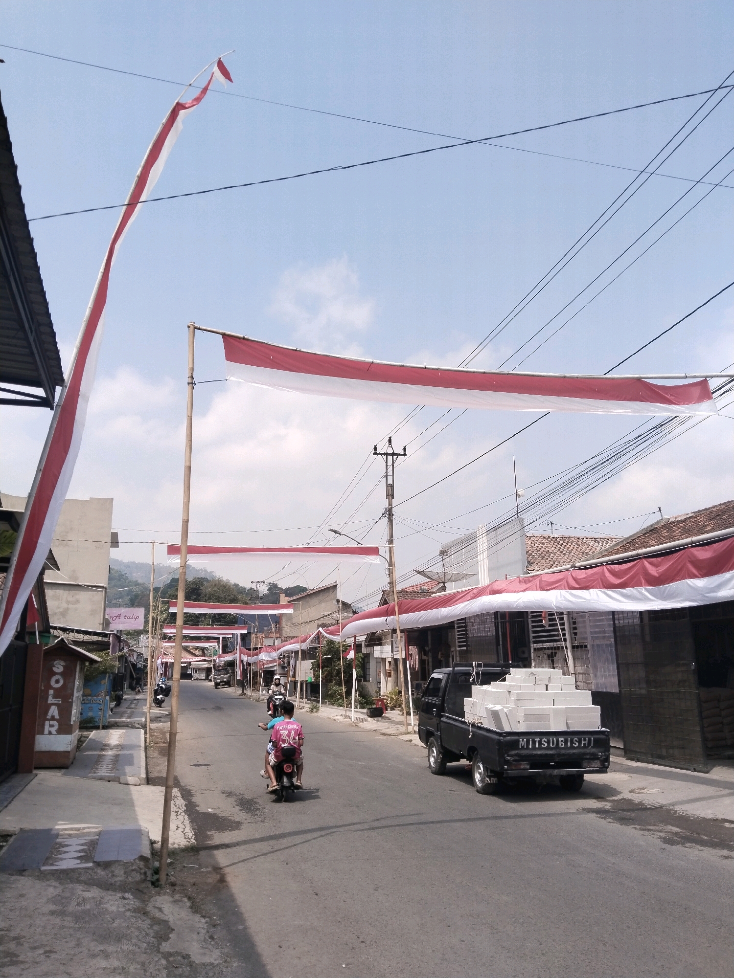The image shows a street scene in Jejeg, Indonesia, likely decorated for a national celebration with red and white Indonesian flags strung across the road. On the left side of the street, there is a building with a sign that reads "SOLAR". Further down the street, two people are riding on a motorcycle. A black Mitsubishi pickup truck is visible in the center of the frame, loaded with white rectangular objects, possibly building materials. Several buildings line the right side of the street, some with awnings. The sky is blue with some clouds, and electrical wires crisscross overhead. The road surface appears to be asphalt.