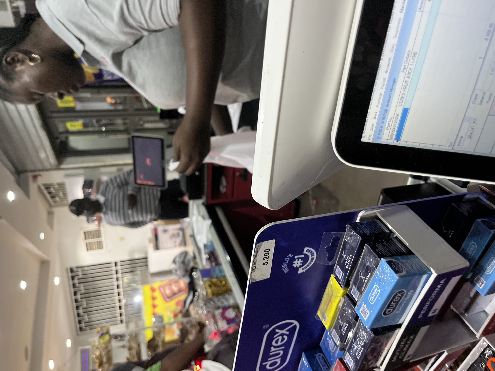 The image shows a cashier at a Dellamond Supermarket in Abuja, Nigeria, ringing up a customer. The point-of-sale system on the screen lists "CERES FRUIT JUICE 1 LITRE." A display of Durex condoms is visible in the foreground, including "PERFORMA," "MUTUAL CLIMAX," and "Extra Safe" varieties. The price sticker on the display shows "5,200."