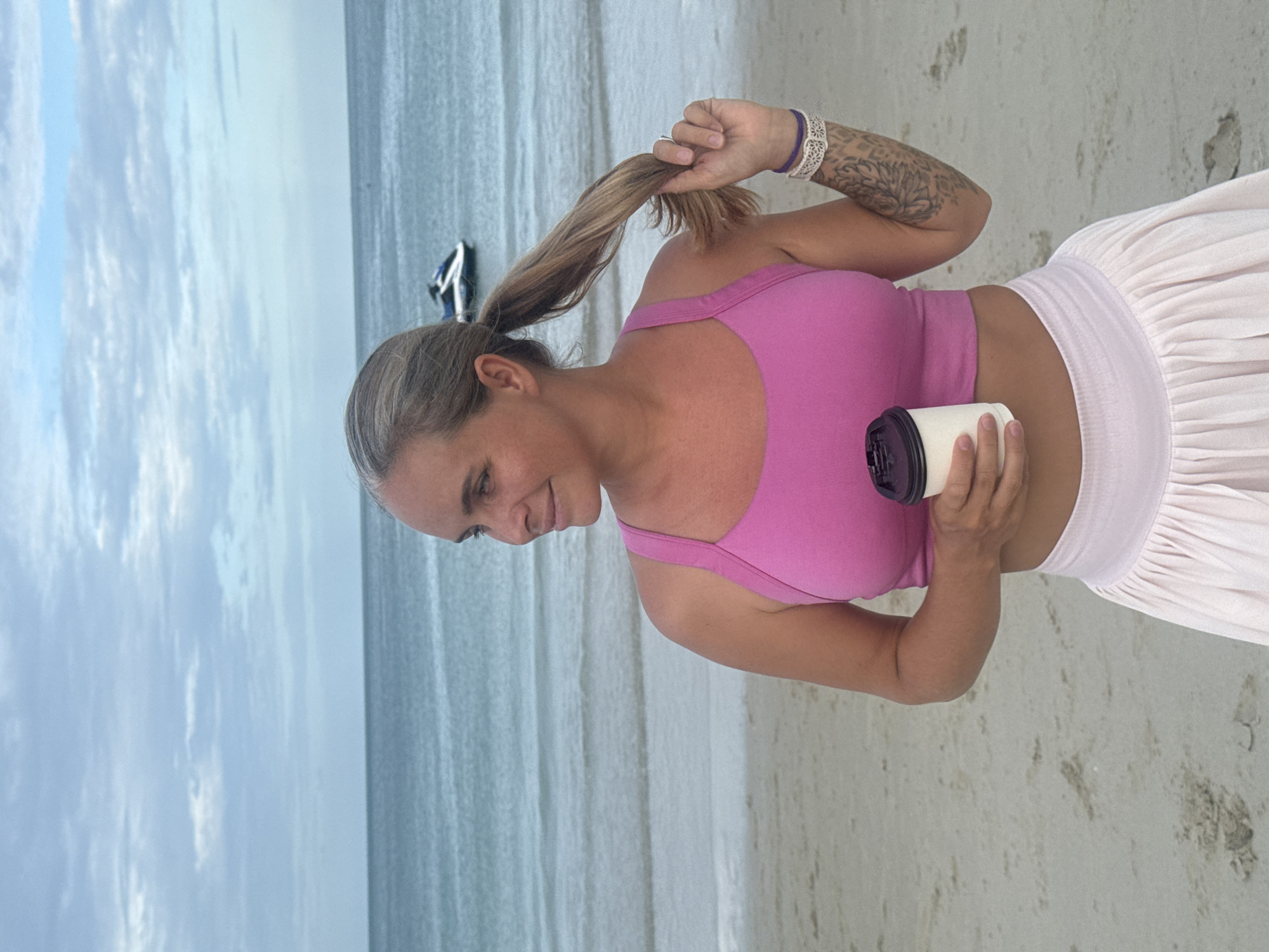 A woman stands on a sandy beach with the ocean and sky in the background. She is wearing a pink sports bra and a light pink skirt. Her hair is pulled back in a ponytail, and she is holding a cup in her right hand while touching her ponytail with her left hand. A jet ski is visible in the water. The scene appears to be in Ko Samui District, Thailand, based on the presence of a jet ski, common in tourist beach destinations there.