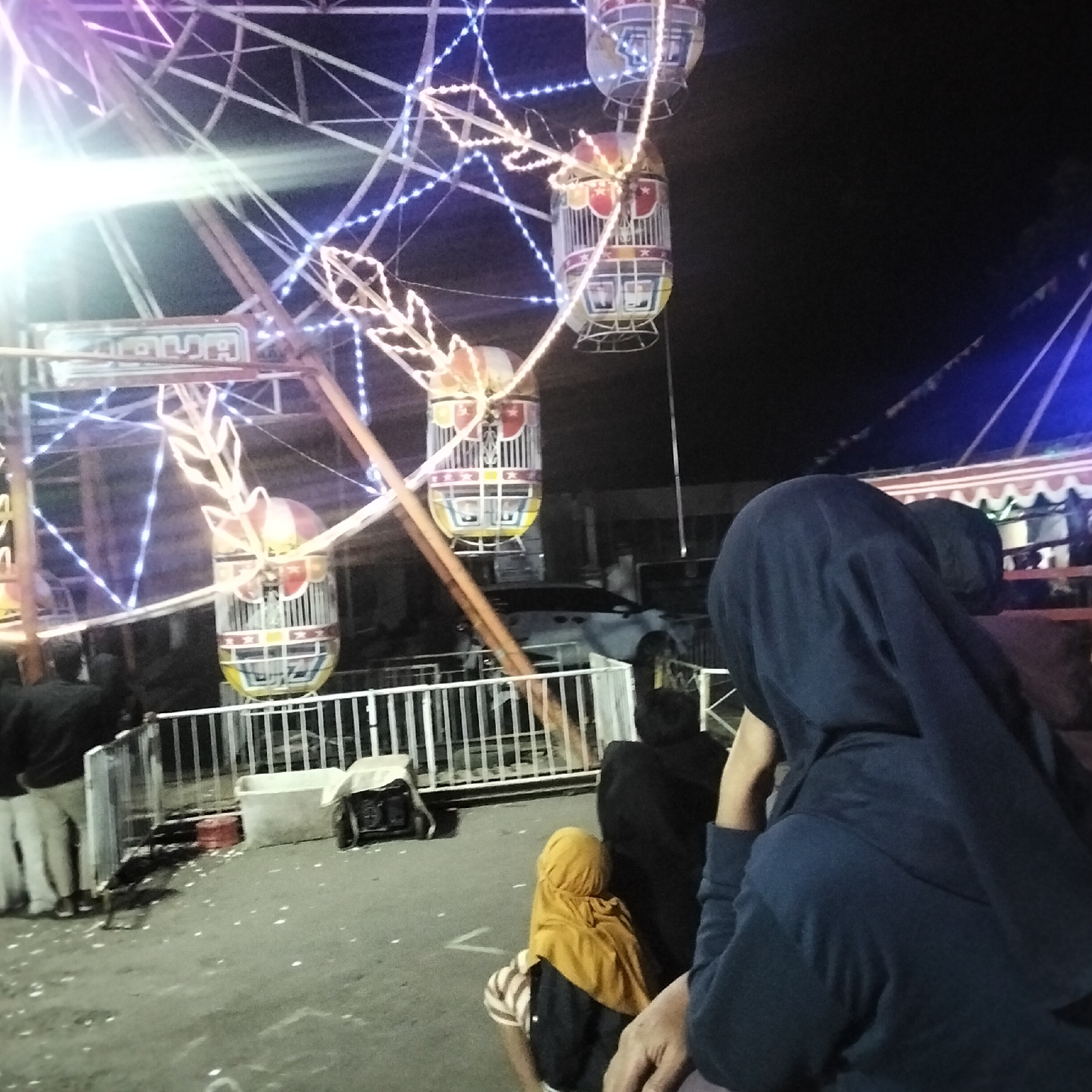 The image depicts a nighttime scene at what appears to be a local carnival or fair in Cisaat, Indonesia. A Ferris wheel, adorned with numerous white and blue lights, dominates the background, with several gondolas visible. In the foreground, a group of people are gathered. On the far left, two individuals are standing, facing away from the camera, wearing dark tops and light-colored pants. Further into the image, a person wearing a yellow hijab and a striped shirt is seated, also facing away. To their right, another person is partially visible, wearing a dark head covering. The ground is paved and appears somewhat uneven, with scattered white debris. A white metal railing separates the gathering area from the operational area of the amusement ride. In the mid-ground, a light-colored car is parked. The overall atmosphere suggests a community event taking place in an open area at night.