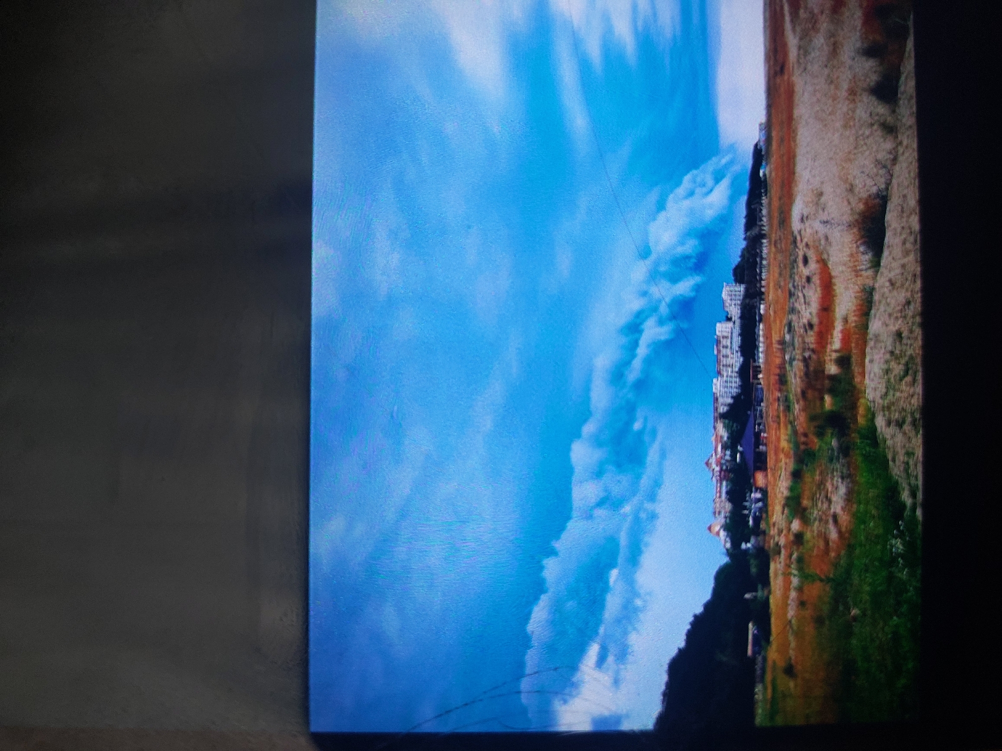 The image shows a landscape scene, likely taken in or near the city of Tsarevo, Bulgaria, judging by the architecture and landscape. The scene is dominated by a bright blue sky with long, stretched-out clouds. To the right of the sky is a shoreline featuring buildings and vegetation. The buildings appear to be residential or hotel structures. The vegetation consists of green grass, brown brush, and dark patches suggestive of trees or forested areas. The image appears to be a photograph of a screen, with the edge of the screen visible on the left. There are visible scratches near the bottom left of the scene. The overall lighting suggests daytime conditions.