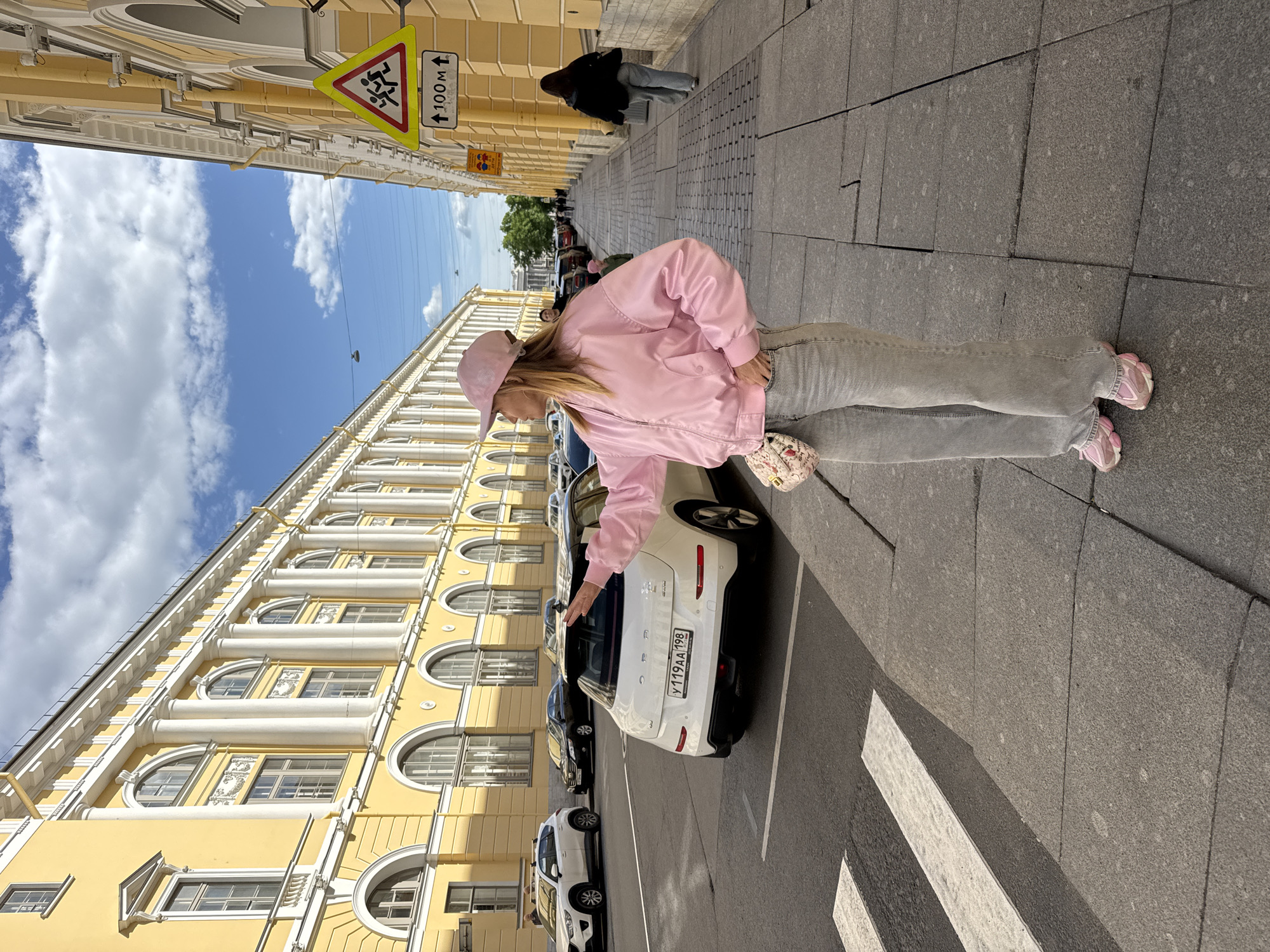 A medium shot shows a woman standing on a sidewalk in Saint Petersburg, Russia, reaching towards a white car. The woman has blond hair and wears a pink baseball cap, a light pink jacket, gray jeans, and pink shoes. She holds a small purse on her left side. The car is parked along the street, with other cars behind it. In the background, there is a tall yellow building with arched windows and white columns, with a blue sky above. A street sign with a "children crossing" symbol and the text "100M" is visible. Other people are walking in the background along the sidewalk. The sidewalk is made of large gray stone blocks, and a pedestrian crosswalk is visible in the foreground. The license plate number is visible on the white car.