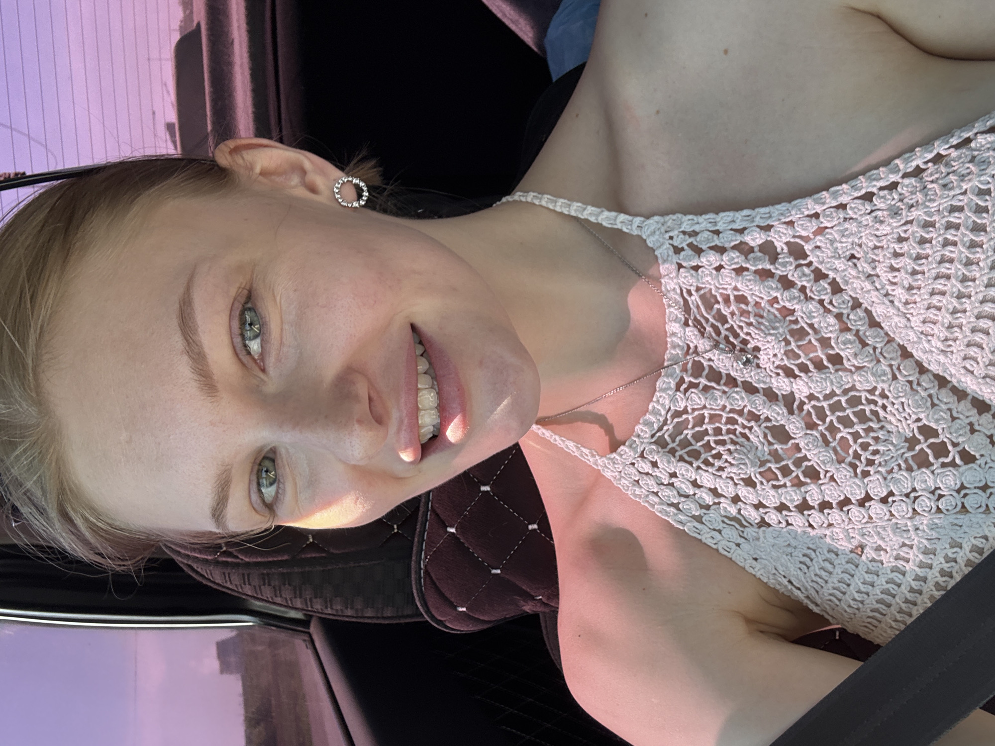 A young woman with light skin, light brown hair, and green eyes is in a car. She is wearing a white crochet top and a silver necklace and earrings. She is smiling, looking at the camera, and appears to be in the passenger seat of the car. The background outside the car window is blurred, showing an urban landscape under a lavender-colored sky. Based on visual information, the location in the city of Perm, Russia, is not discernible.