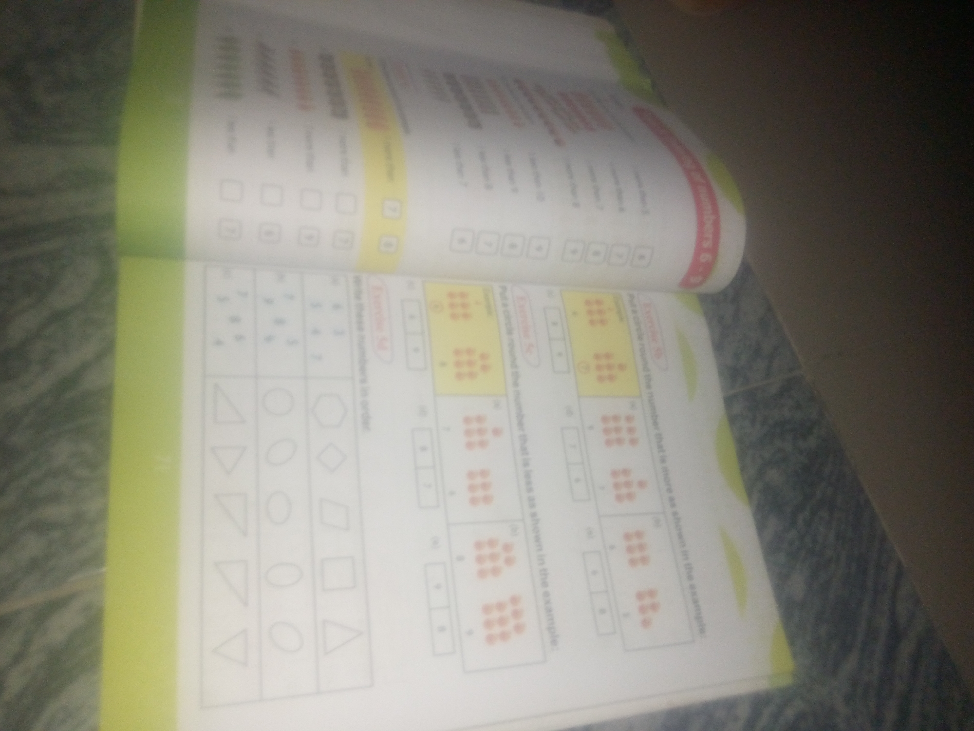 The image shows an open book on a textured surface. The book is a children's workbook with various exercises. One page features a grid of geometric shapes, including triangles, circles, hexagons, diamonds, and squares. Another section of the page contains exercises with numbers and dots, likely for counting or comparison. The workbook has a yellow border on the left side and a red curved design on the right. The overall scene appears to be a close-up, angled shot of the workbook. There is no indication of a specific city or location like Jauro Yinu, Nigeria.