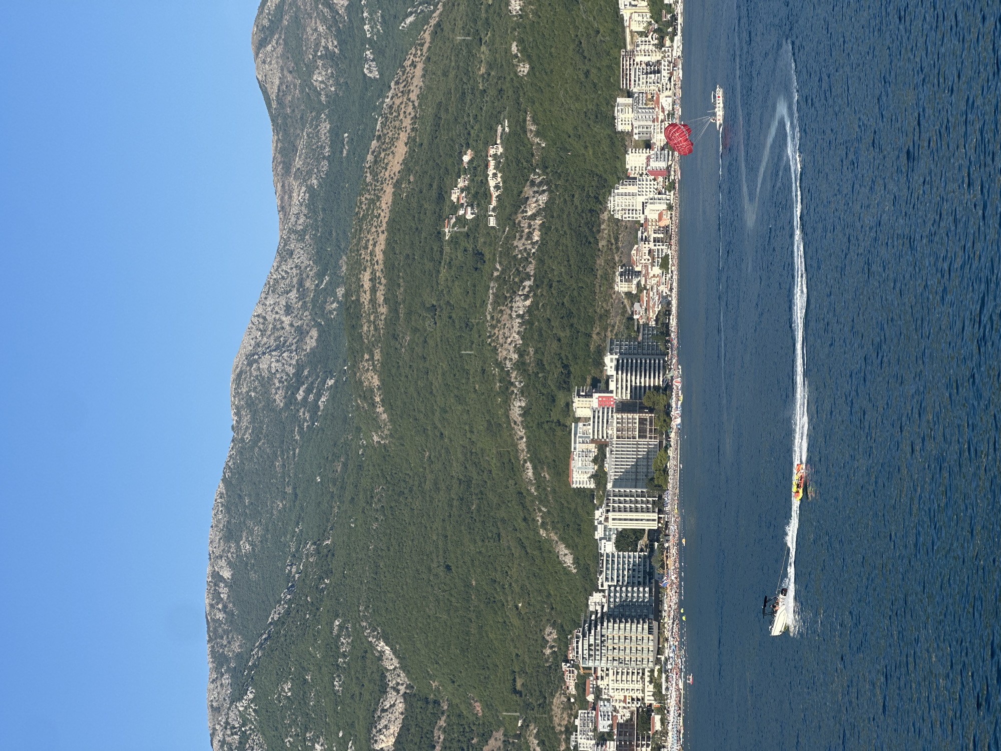 This image depicts a coastal scene in Boreti, Montenegro, featuring a wide expanse of deep blue water in the foreground. A white speedboat is actively engaged in towing a yellow inflatable tube with at least two individuals on it, leaving a prominent white wake in its path. To the right, a parasailor, attached to a red parachute, is being towed by another boat, also creating a swirling white trail on the water. In the mid-ground, a densely populated beach is visible, lined with numerous sunbathers and umbrellas, indicating a popular tourist destination. Behind the beach, a modern cityscape with numerous multi-story buildings and hotels stretches along the coastline. Dominating the background are lush green mountains, partially covered with trees and rocky outcrops, under a clear blue sky. The overall scene captures a vibrant summer day with recreational activities on the water and a busy resort town.