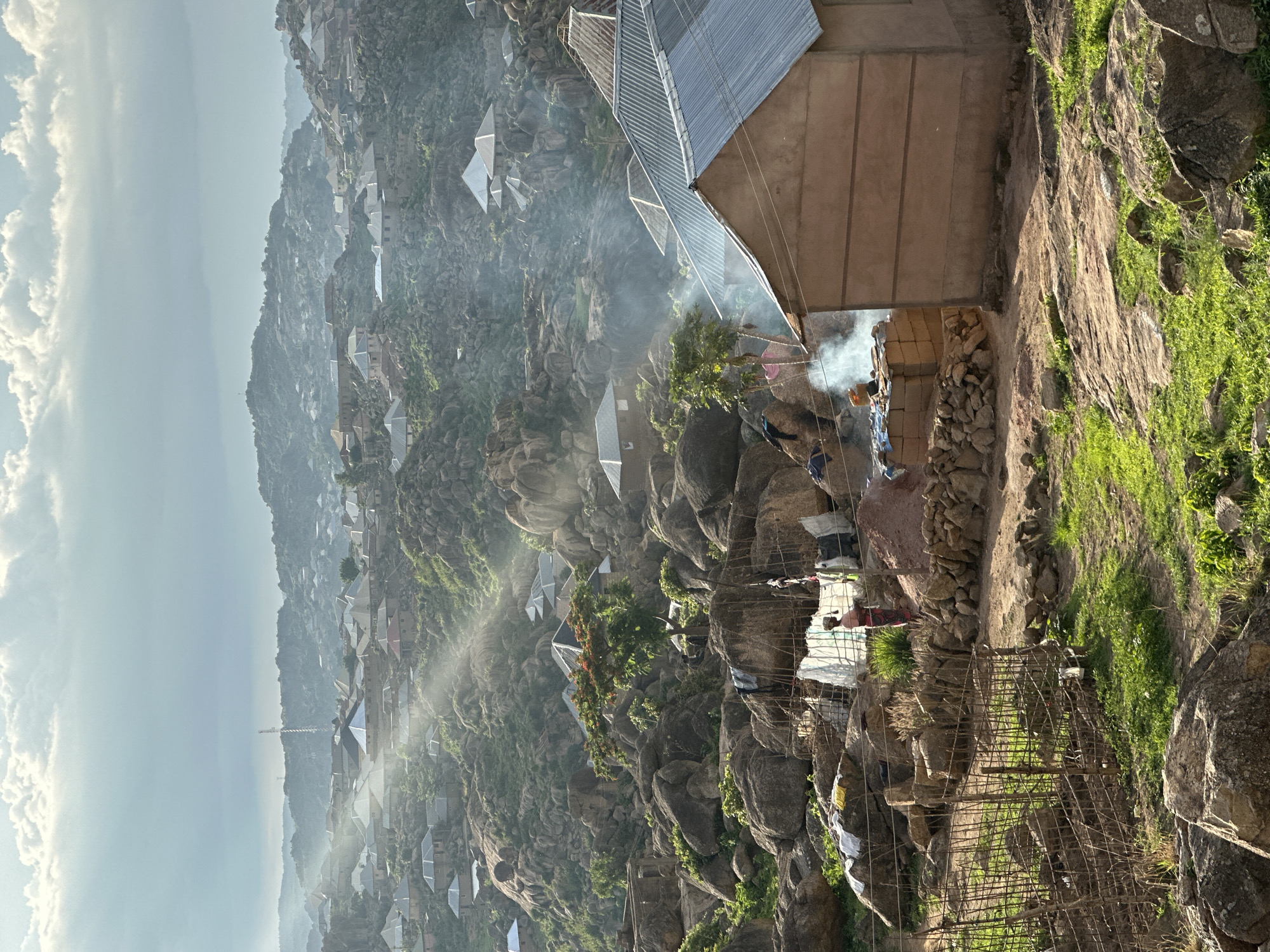 The image depicts a densely populated, rocky urban landscape in Jos, Nigeria, under an overcast sky with scattered sunlight. Numerous single-story residences with corrugated metal roofs are integrated into a terrain dominated by large granite boulders and sparse green vegetation.
In the foreground right, a light brown house with a grey corrugated metal roof is prominent. Behind this structure, white smoke ascends from a ground-level fire, where two individuals are observed; one directly tending the fire, another further back. To the left, a section of green ground is enclosed by a rudimentary fence of sticks and wire.
In the mid-ground, a person in red patterned clothing stands near a pole-and-mesh structure used for drying clothes. A pile of reddish-brown earth is adjacent to this area. Throughout the scene, clothes are draped on rocks for drying.
The background shows the settlement extending across multiple hills, featuring similar residential structures. Sunbeams penetrate the clouds, illuminating parts of the landscape and rooftops through atmospheric haze. A tall communication tower is visible on a distant hill. Multiple plumes of smoke are observed across the scene, indicating various fire-related activities.